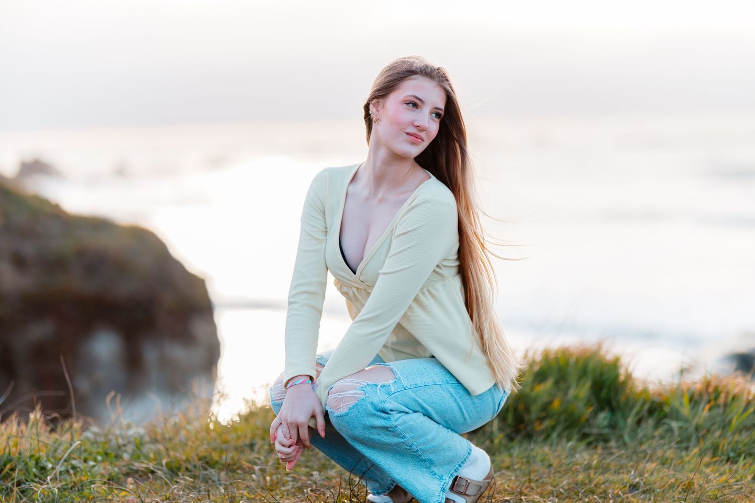 Young woman with long hair in light cardigan and jeans crouching on coastal cliff.