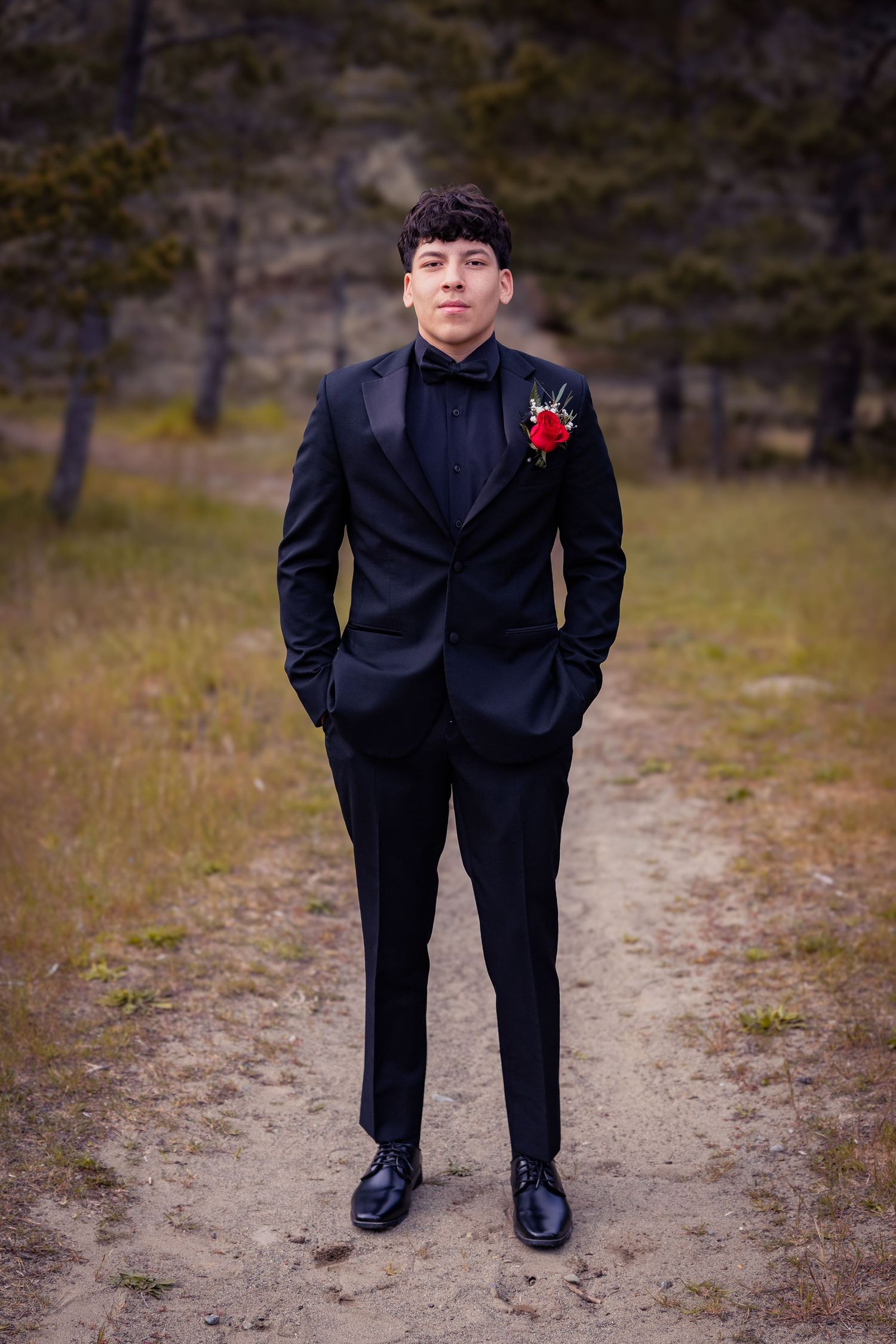 Young man in black tuxedo with red rose boutonniere poses confidently on sandy path for prom portrait outdoors.