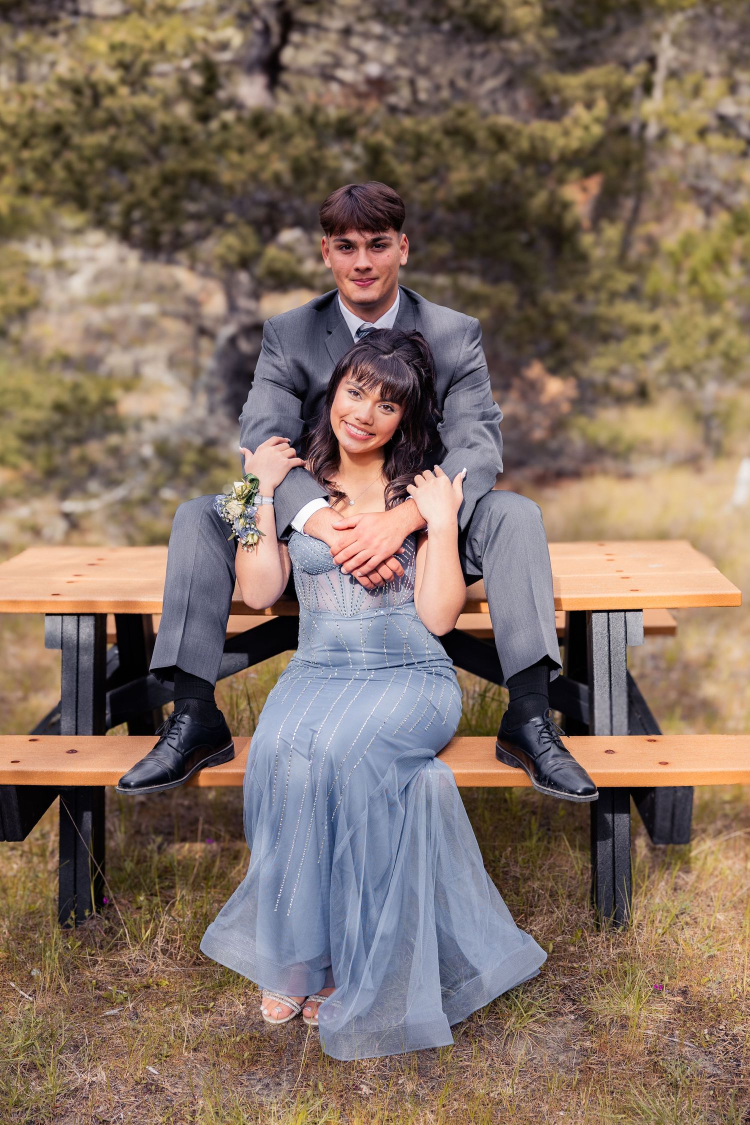 A young couple in prom attire poses at a picnic table outdoors, she in a blue gown and he in a gray suit.