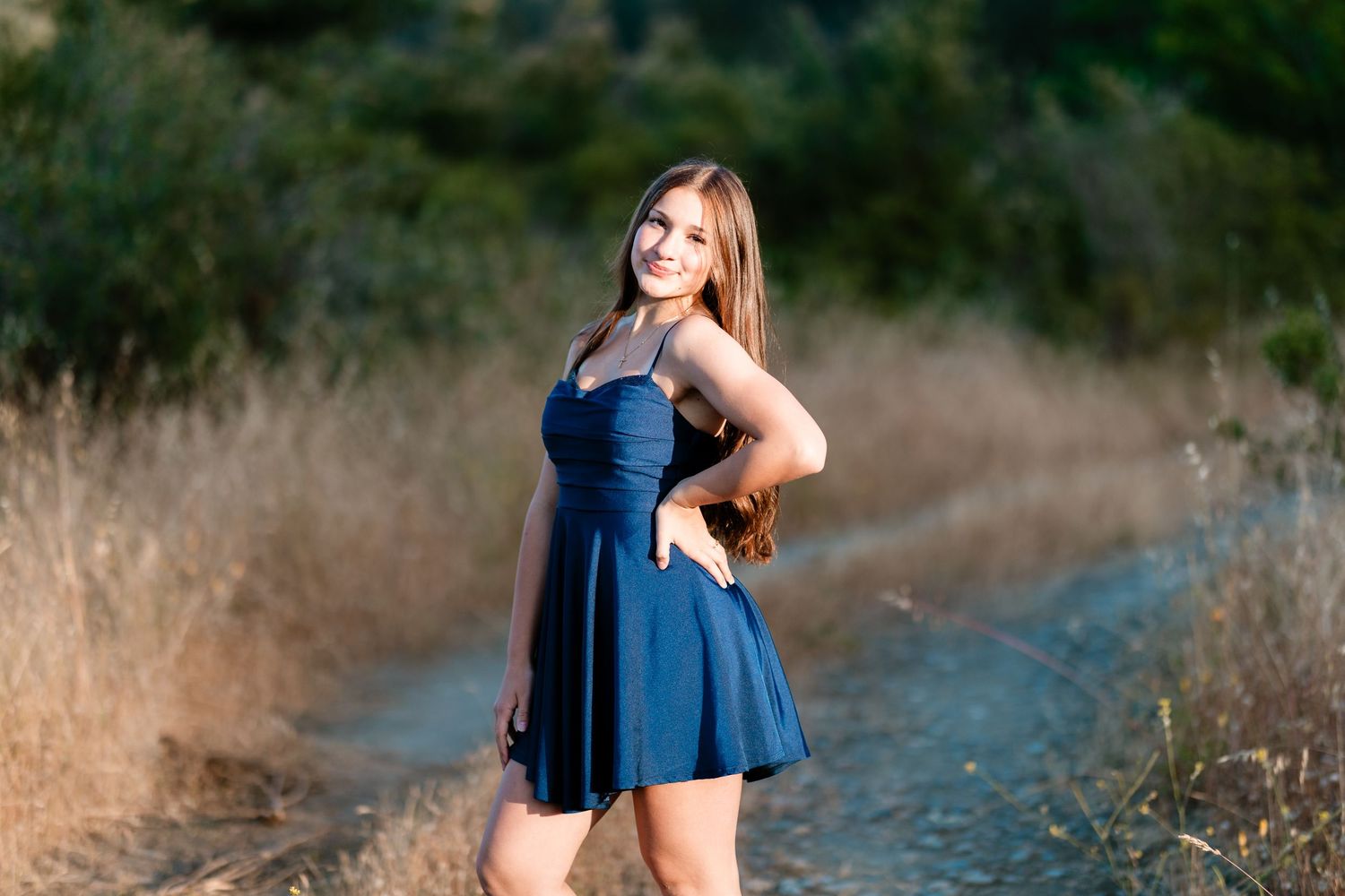 Young woman in navy blue dress posing on a dirt path surrounded by golden grass and green trees.