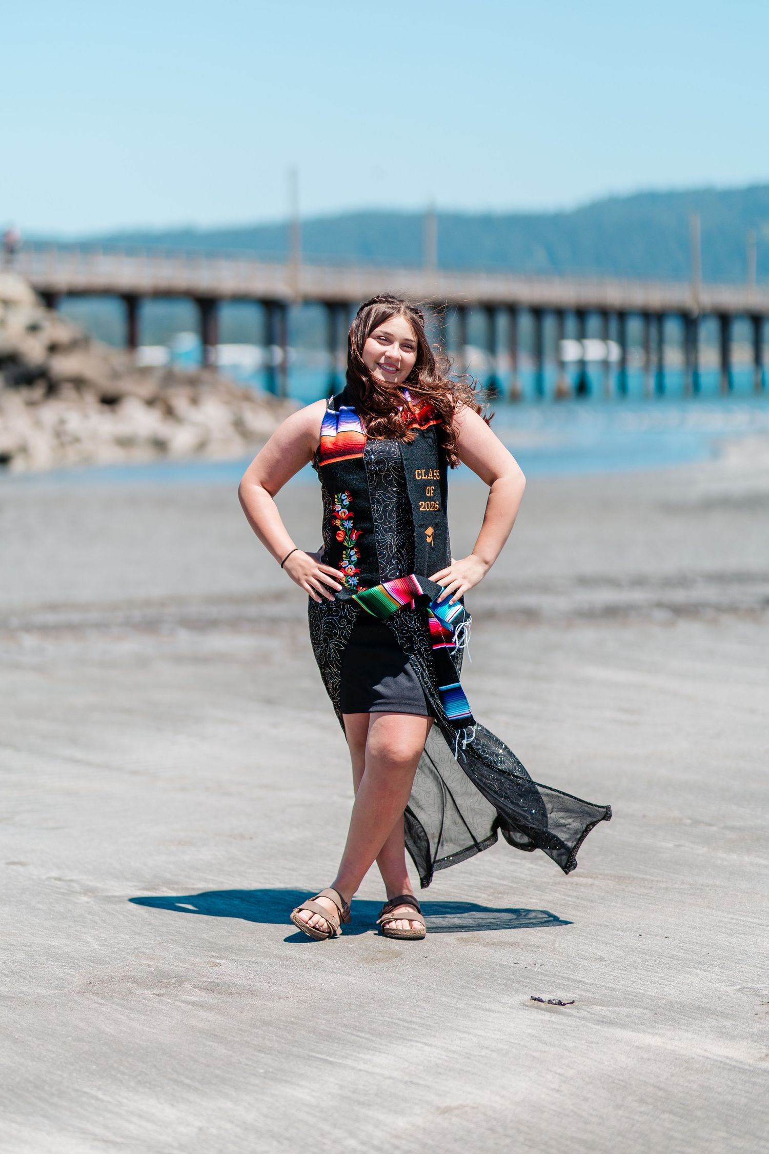Young woman in graduation stole poses confidently on a sunny beach boardwalk near a pier.