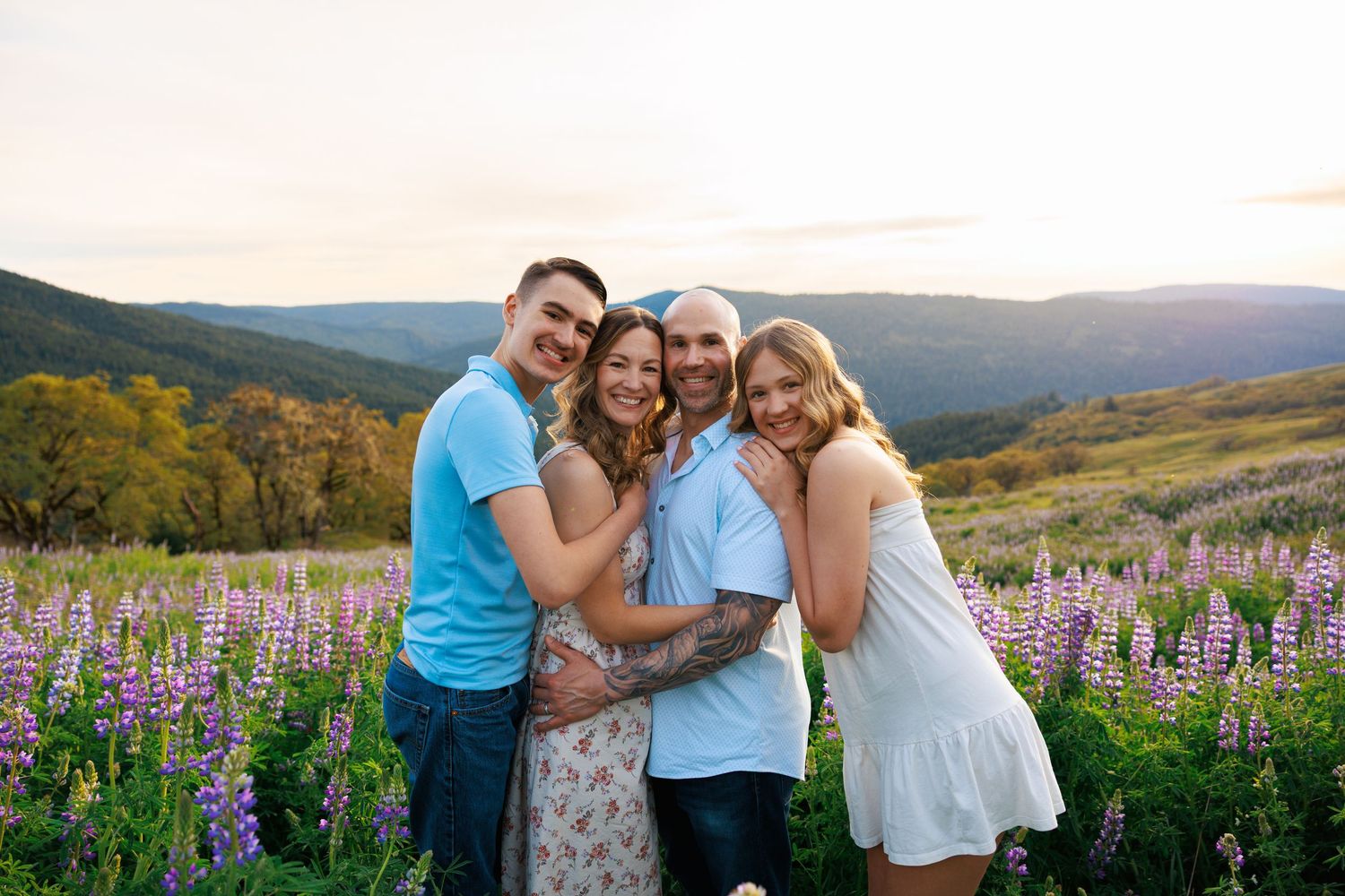 Smiling family of four embracing in blooming purple lupine field at mountain sunset.