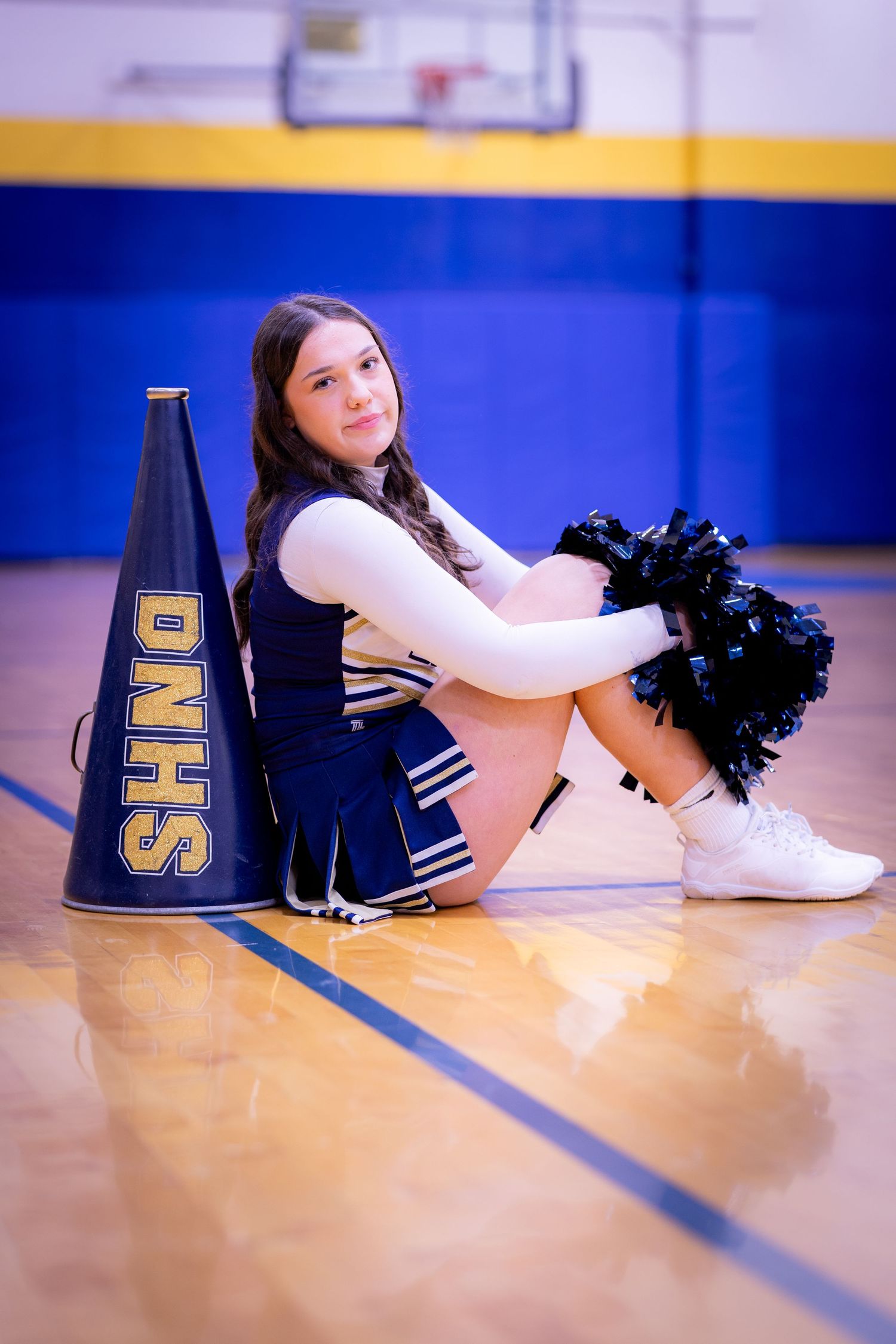 DNHS cheerleader sits on gym floor with pom-poms and navy megaphone.