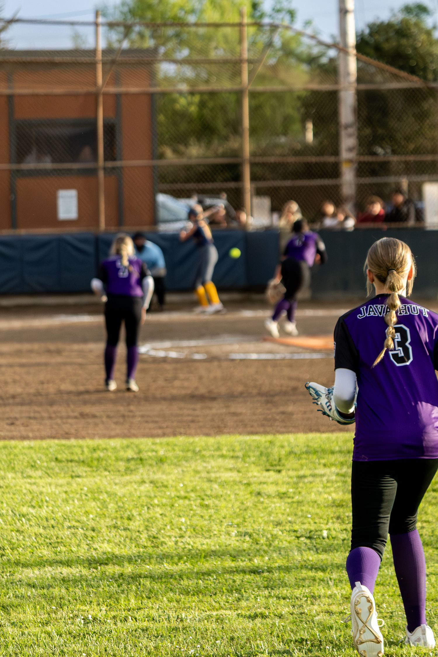 Female softball player wearing number 3 purple jersey watches pitcher throw during an outdoor game.