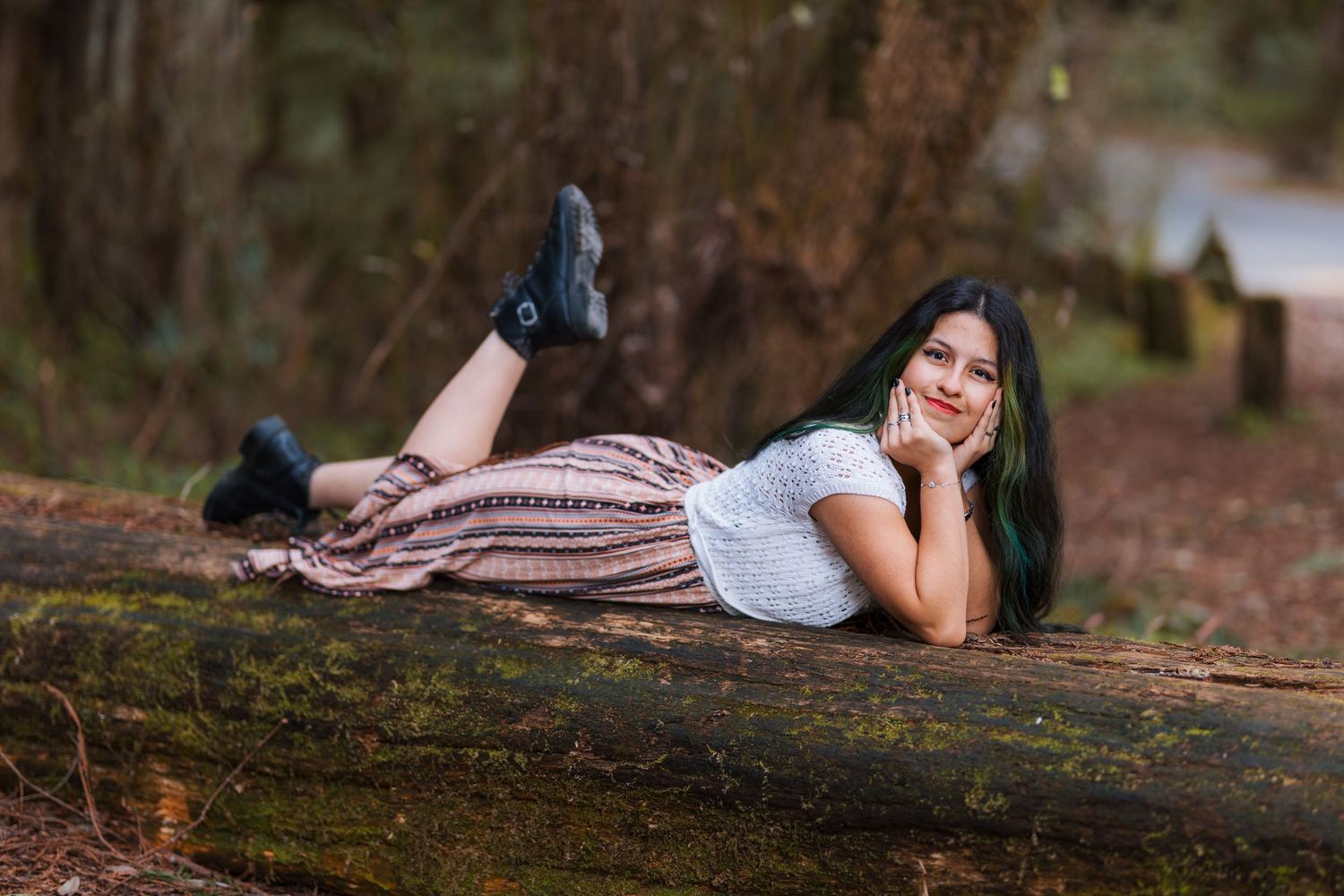 Young woman in striped maxi skirt and black boots poses lying on a mossy log in the forest.