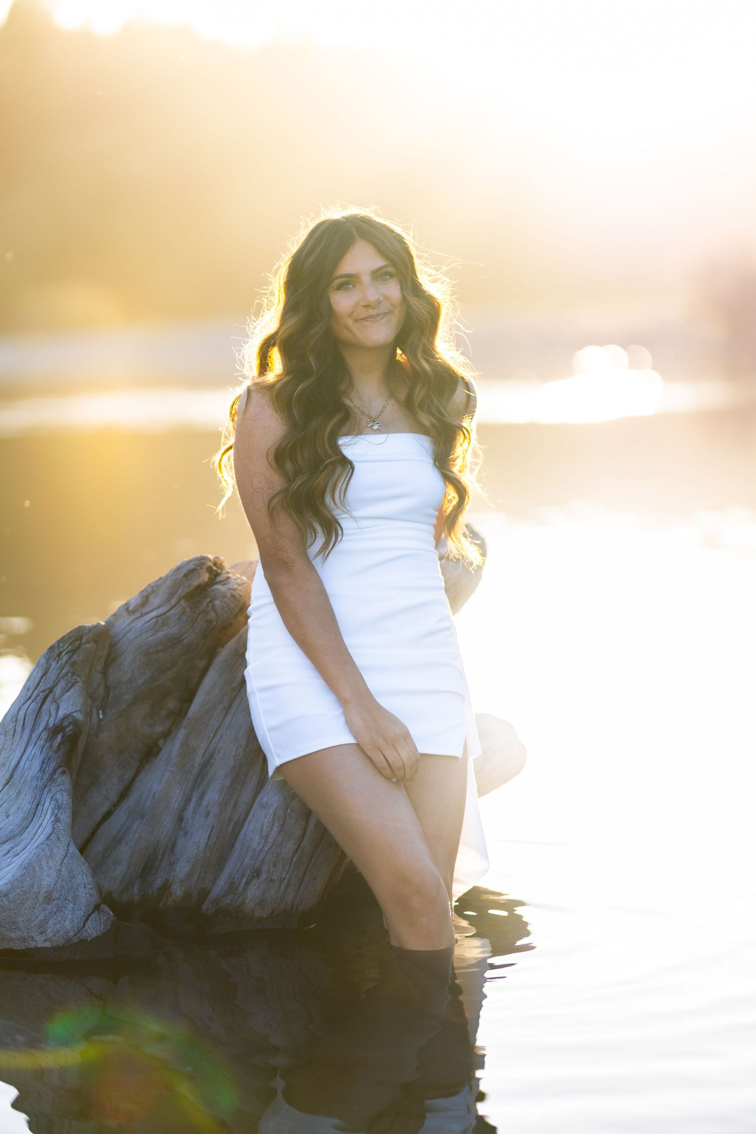Young woman in white dress sitting on driftwood in water with golden sunset light behind her.