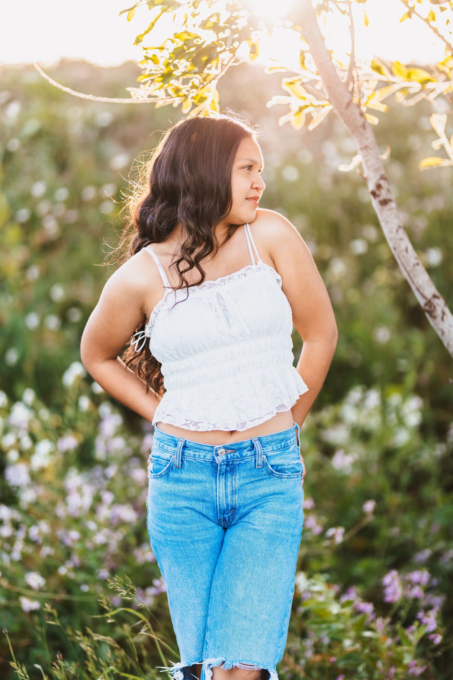 Young woman in white crop top and blue jeans standing in a wildflower field with golden sunlight.