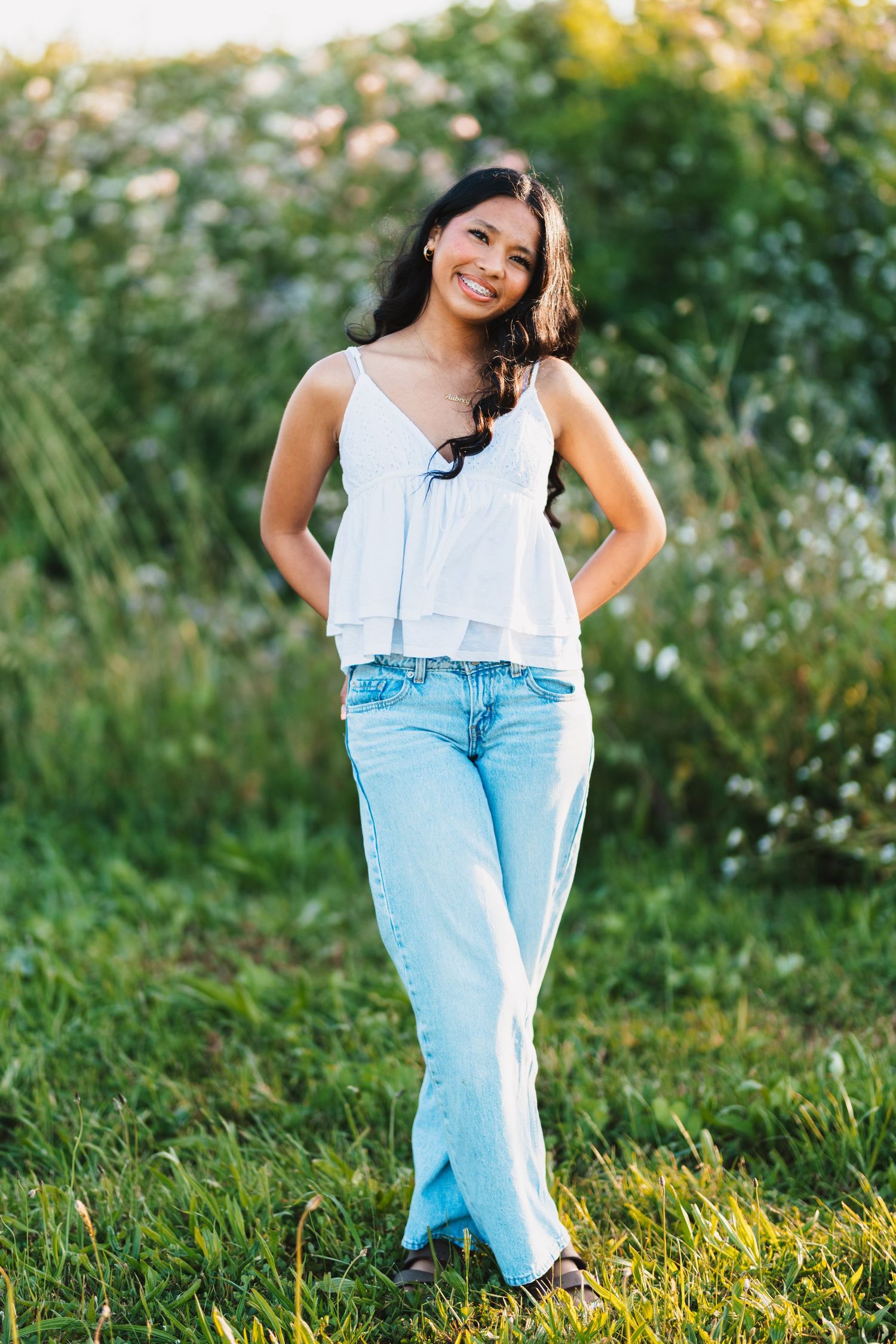 Young woman smiling in white tank top and light blue jeans standing outdoors in a green garden setting.
