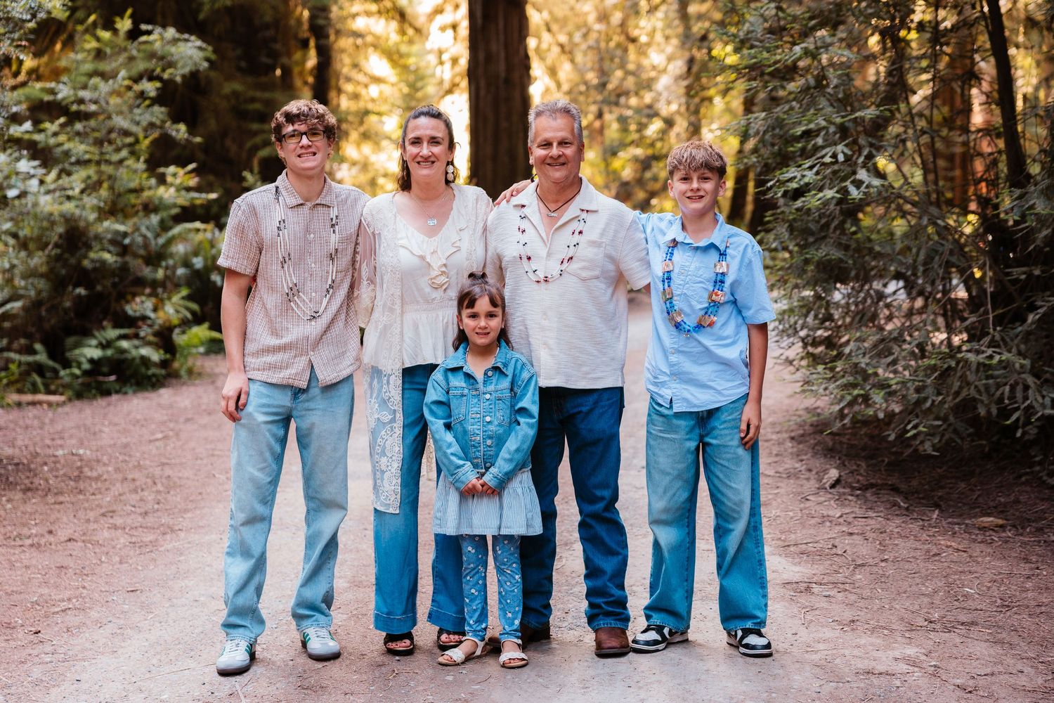 Family of five smiling together on a forest path surrounded by tall redwood trees.