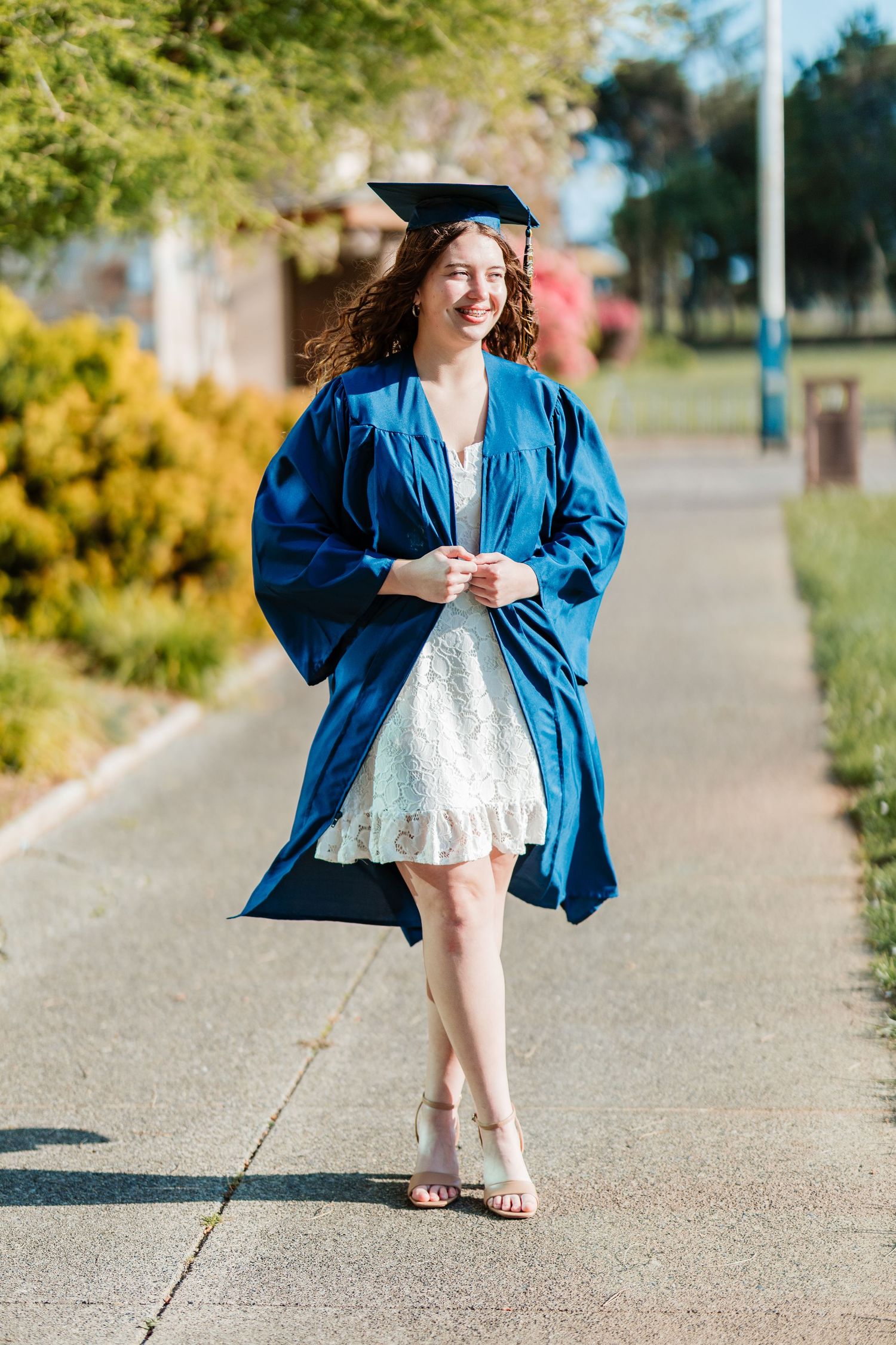 Smiling graduate in blue cap and gown over white dress walking outdoors on a sunny day.