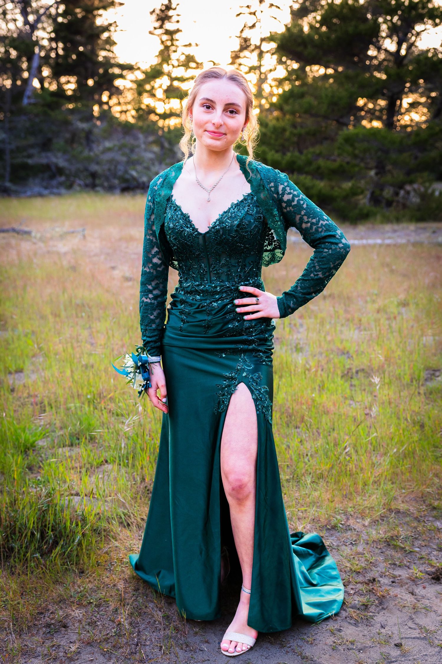 Young woman in elegant green lace long-sleeve prom dress with slit, posing outdoors at golden hour.