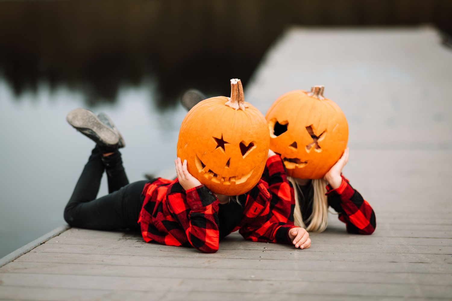 Two people lying on dock holding carved jack-o-lanterns over their faces near water.