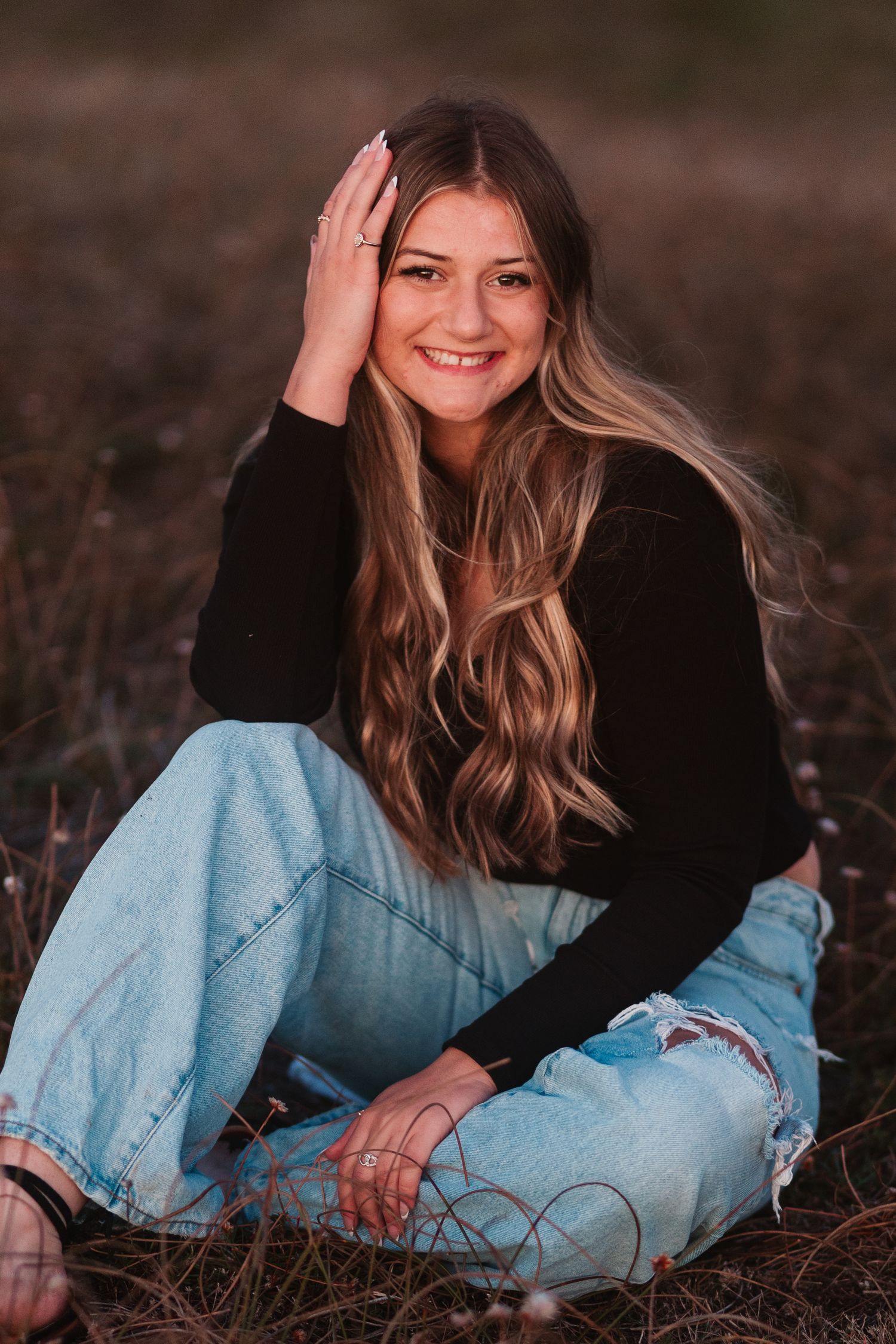 Smiling blonde teen girl in black top and ripped jeans sitting in a field during golden hour.