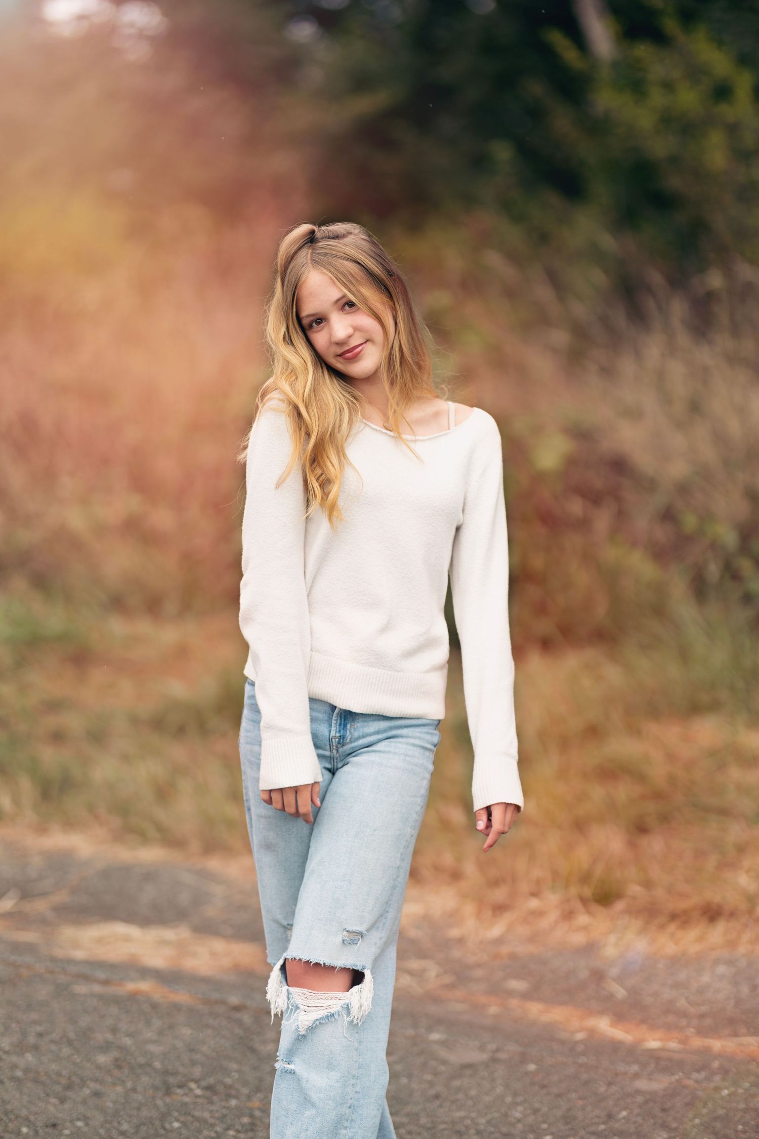Blonde teenage girl in white sweater and ripped jeans posing on outdoor path with autumn background.
