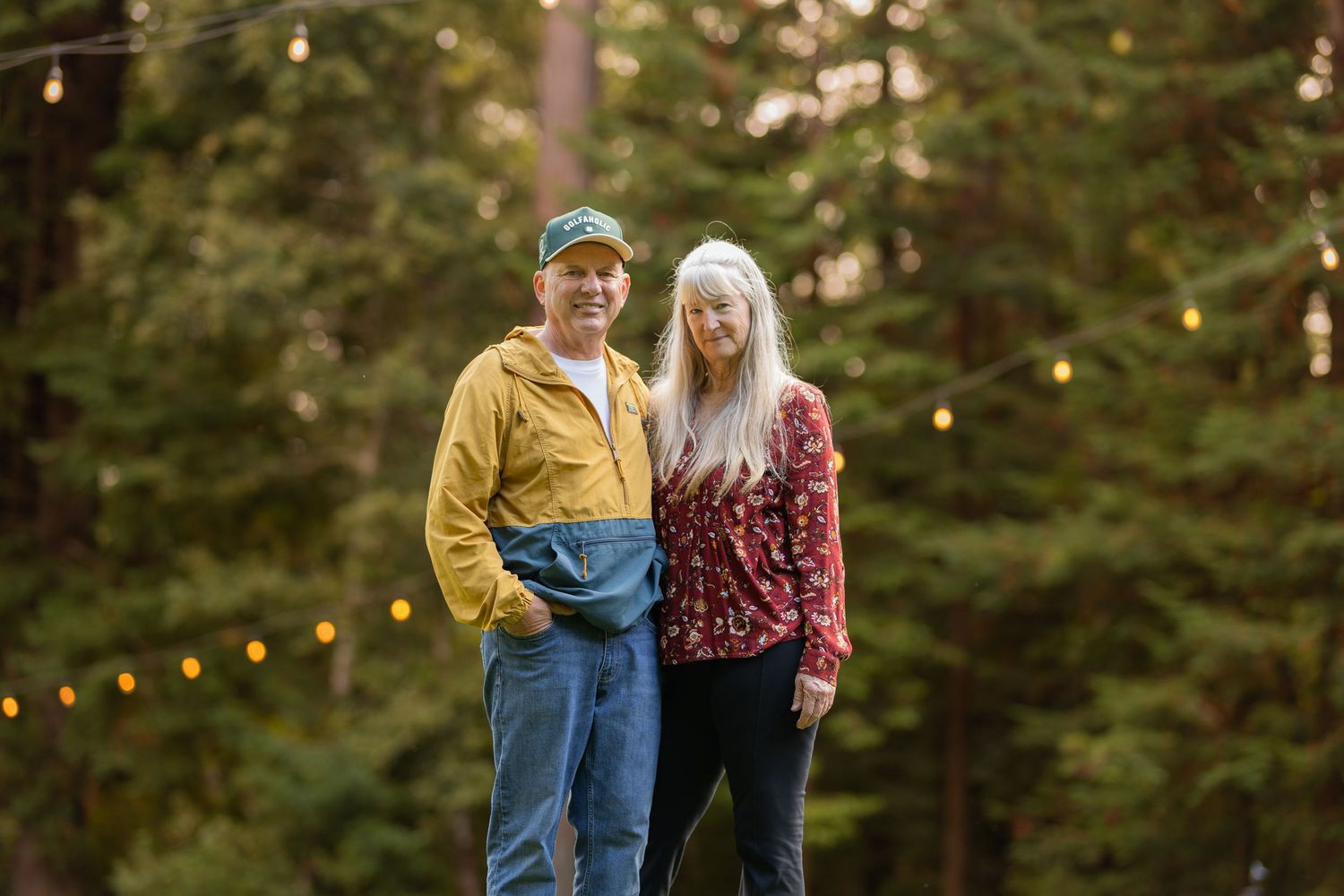 Couple standing together among tall trees with string lights, man in yellow jacket and woman in red floral top.