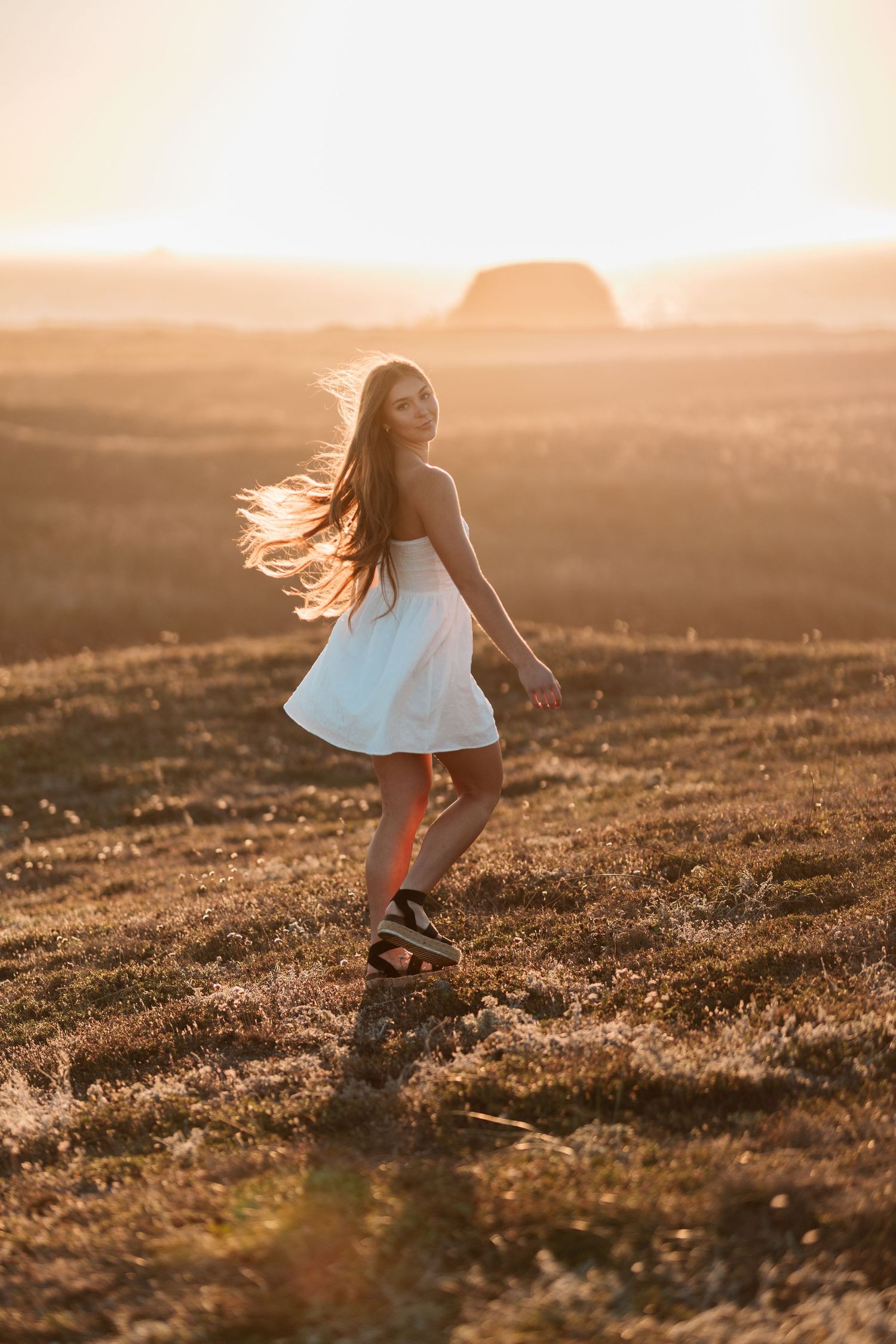 Young woman in a white dress twirling in a golden sunset field with wind in her long dark hair.