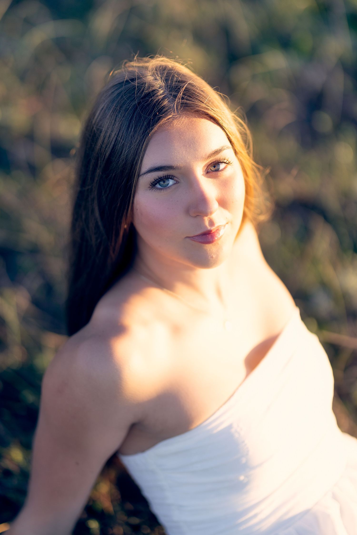 Young woman with long dark hair in a white dress posing in a sunlit field at golden hour.