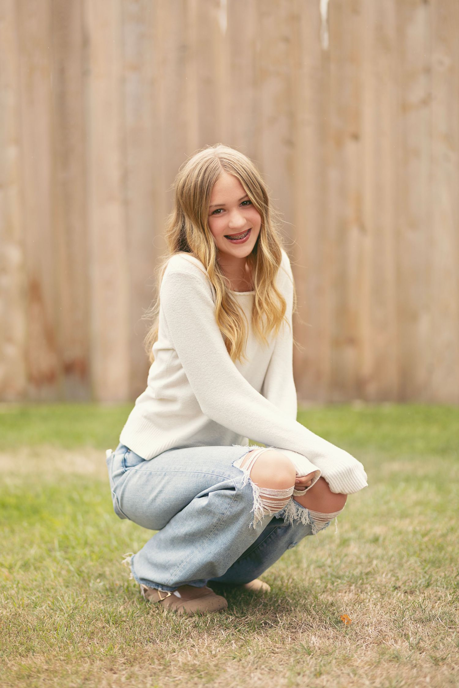 Smiling blonde girl in white sweater and ripped jeans crouching near wooden fence.