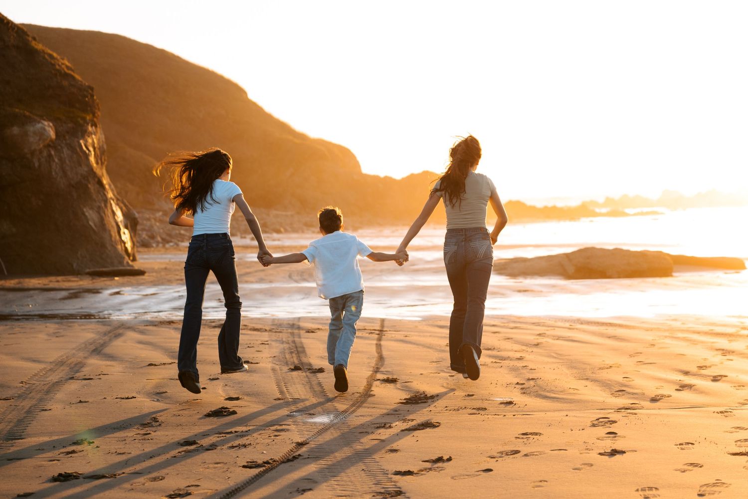 Two women and a child jumping together while holding hands on a beach at warm sunset.
