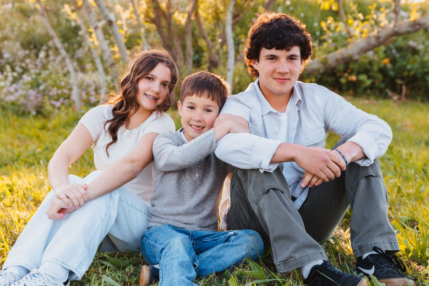Three siblings sitting together on grass, smiling, with blooming trees in the background during golden hour.