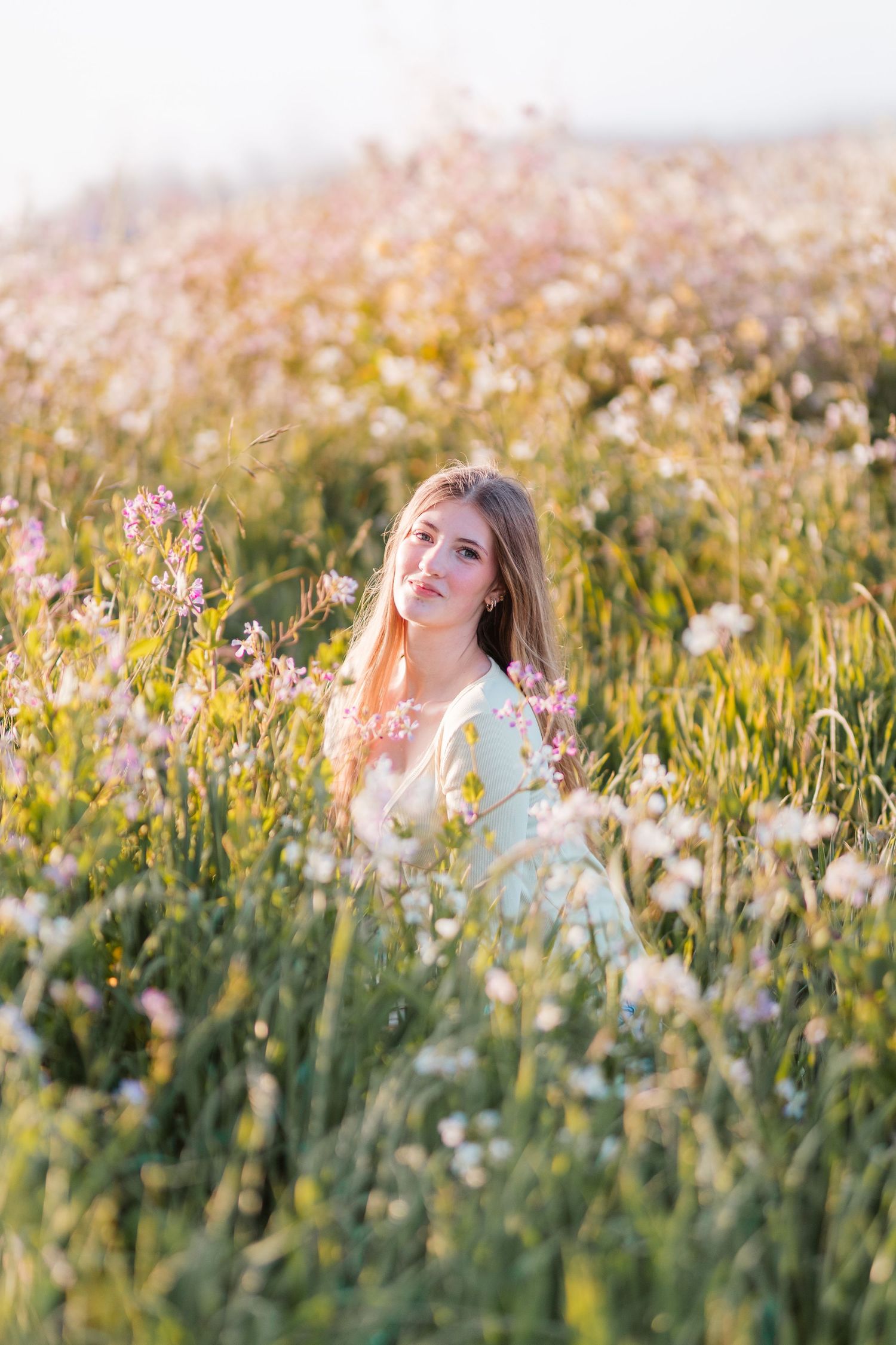 Young woman in light blue dress sits among wildflowers in a sunlit meadow at golden hour.