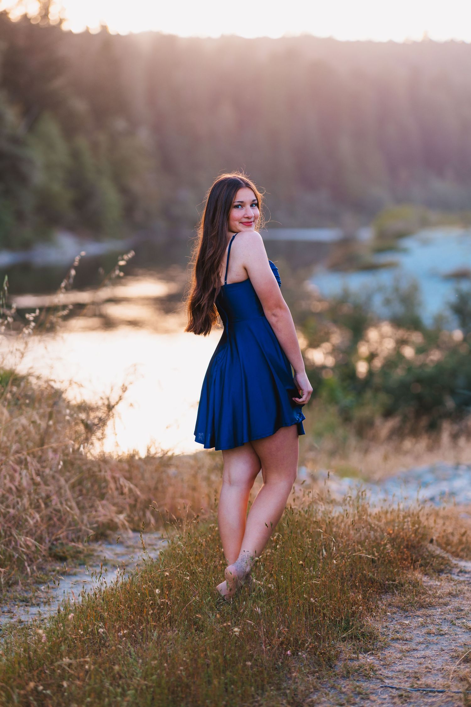 Young woman in blue dress standing on a sunlit path by a river at golden hour, smiling over her shoulder.