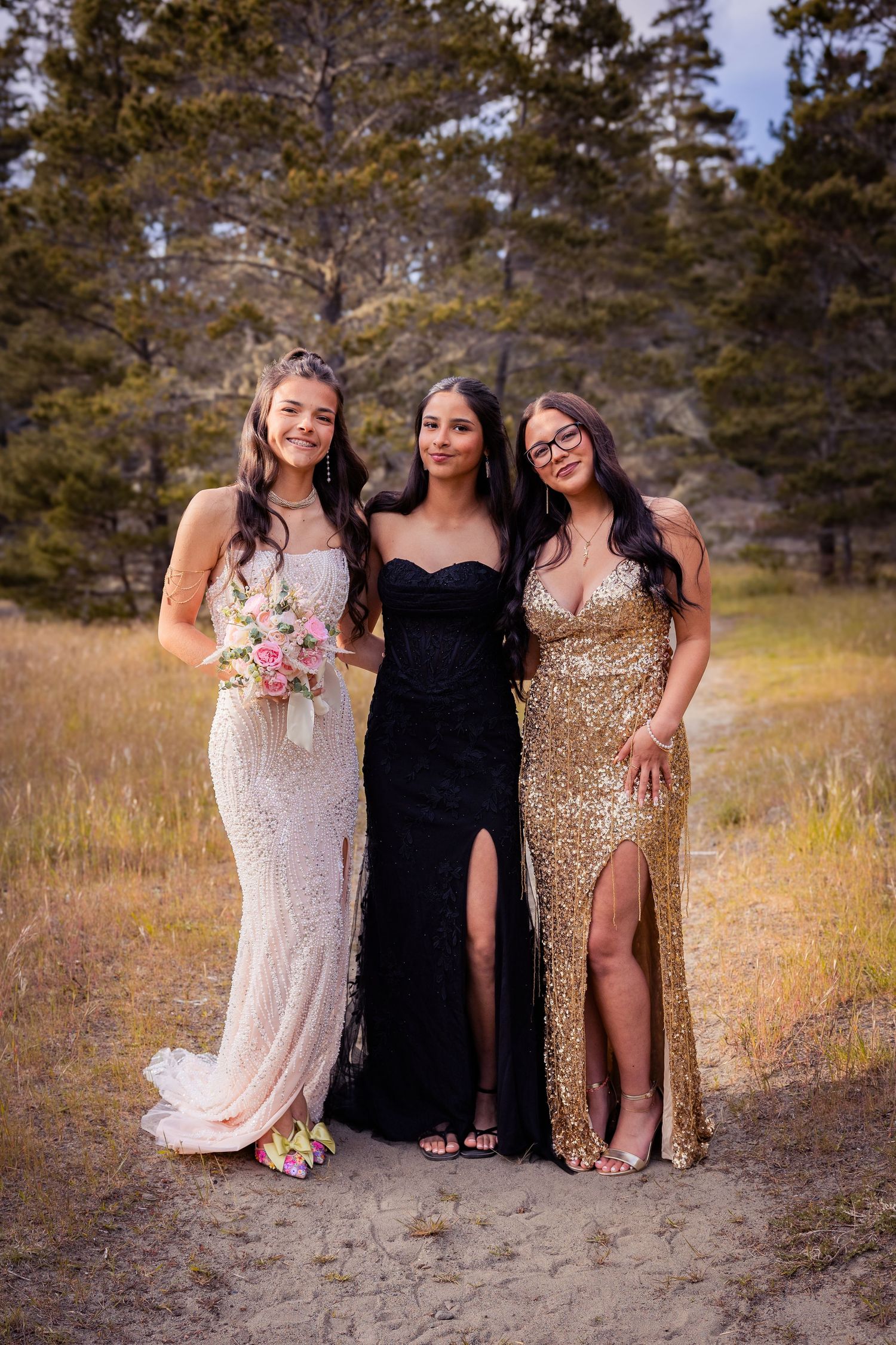 Three young women pose in elegant prom gowns outdoors, wearing white, black, and gold sequin dresses.
