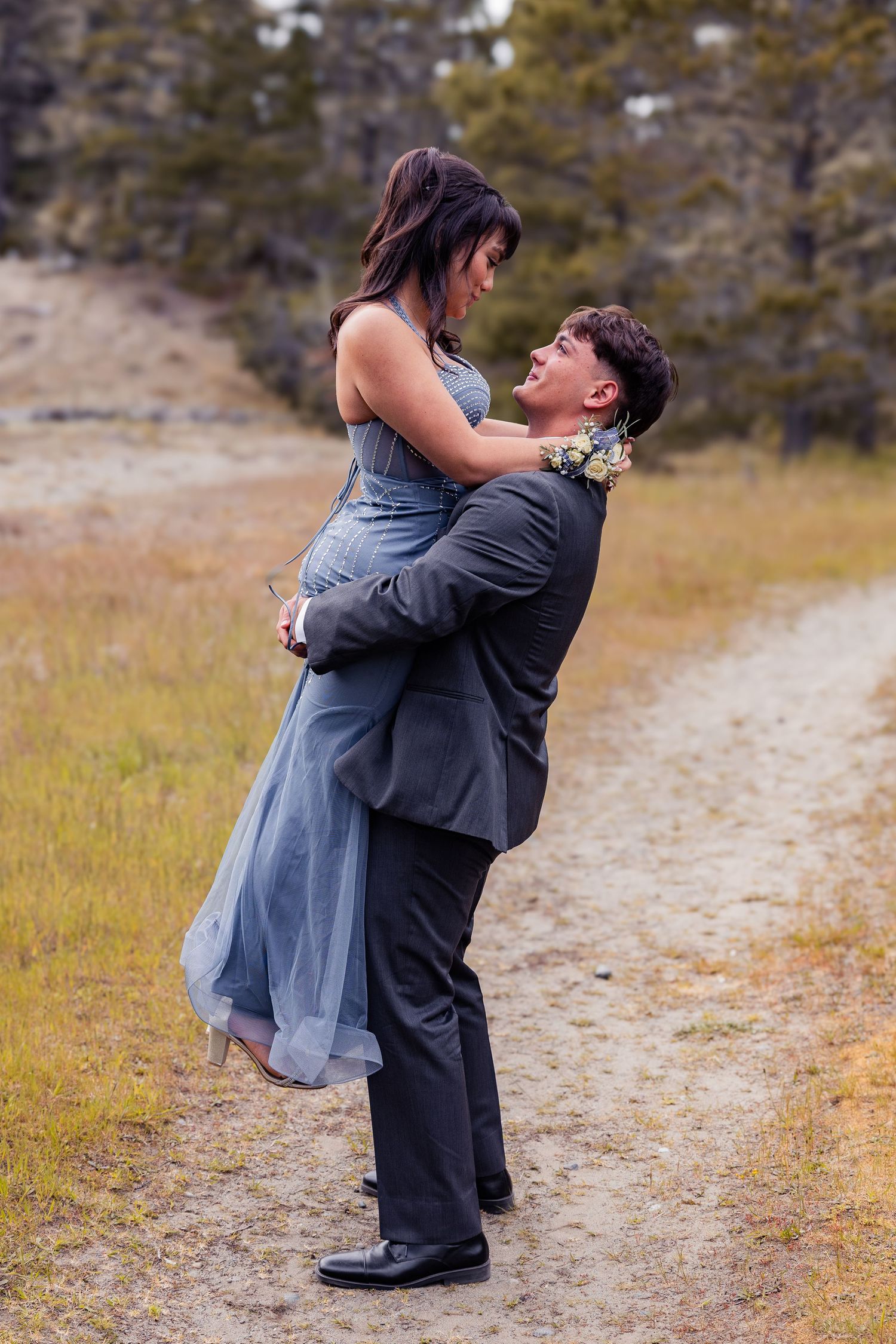 A young man in a gray suit lifts a woman in a blue gown on a dirt path surrounded by golden grass.