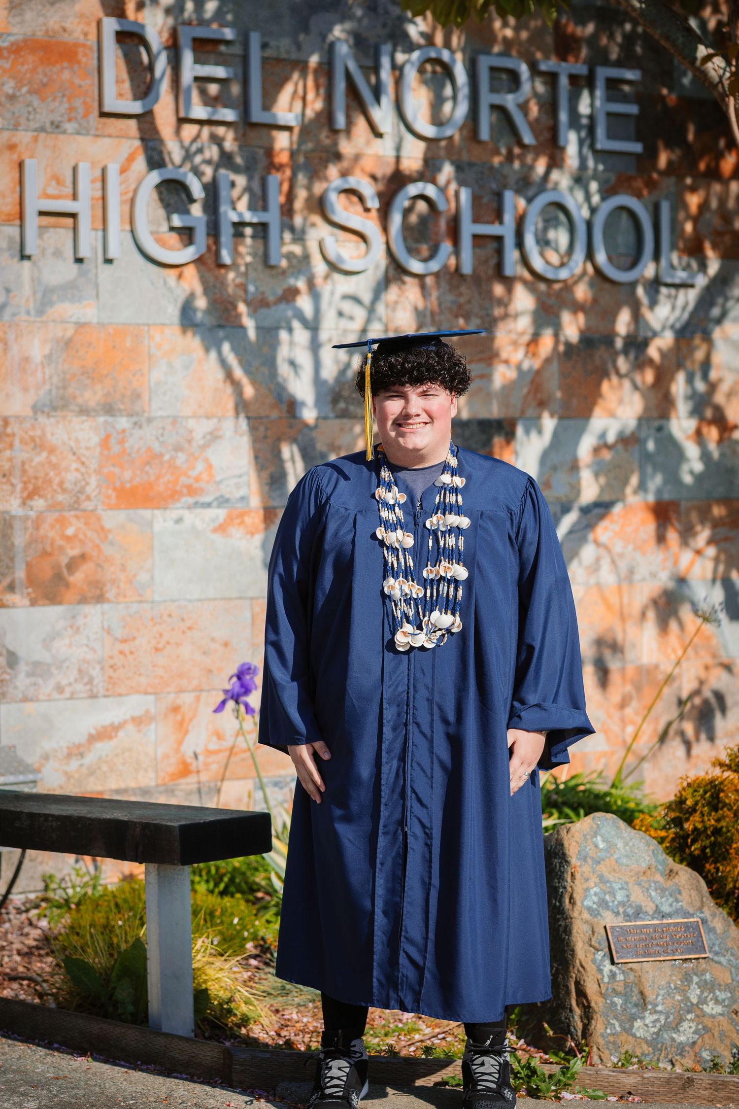 A smiling graduate in blue cap and gown stands proudly in front of Del Norte High School sign.