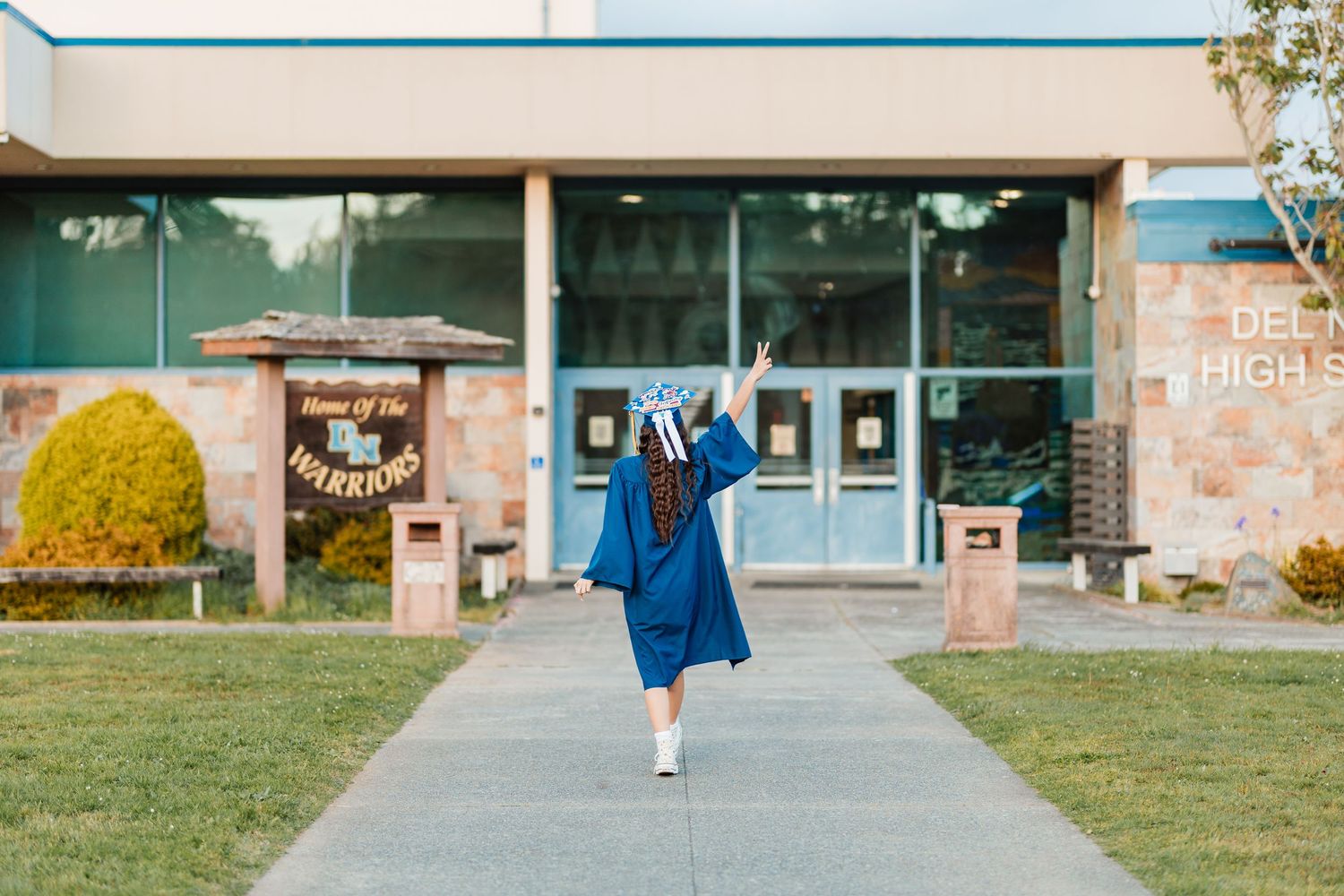 Graduate in blue cap and gown walks toward Del High school entrance, raising hand in celebration.