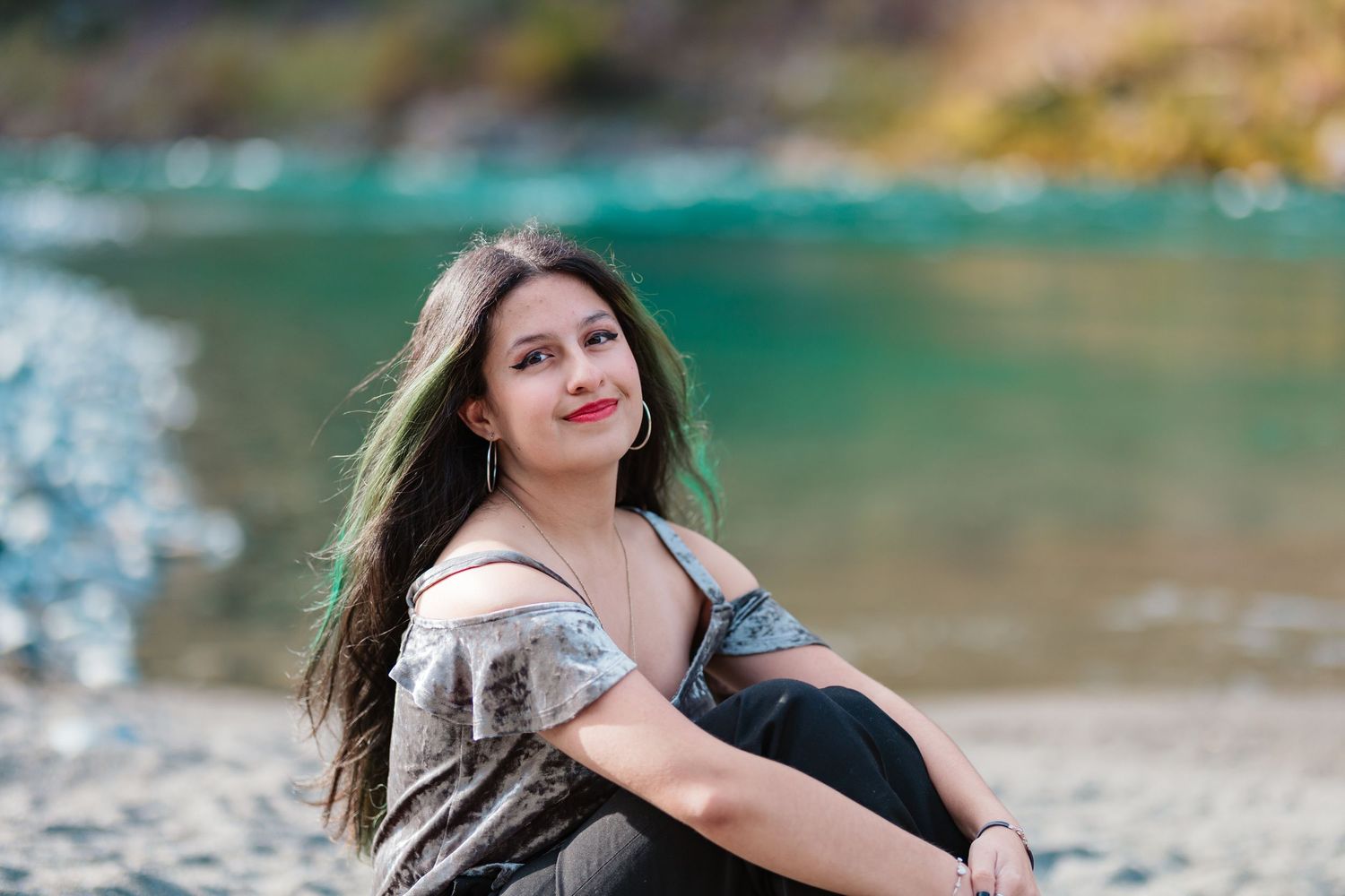Young woman with long dark hair and red lips sitting gracefully by a turquoise river.
