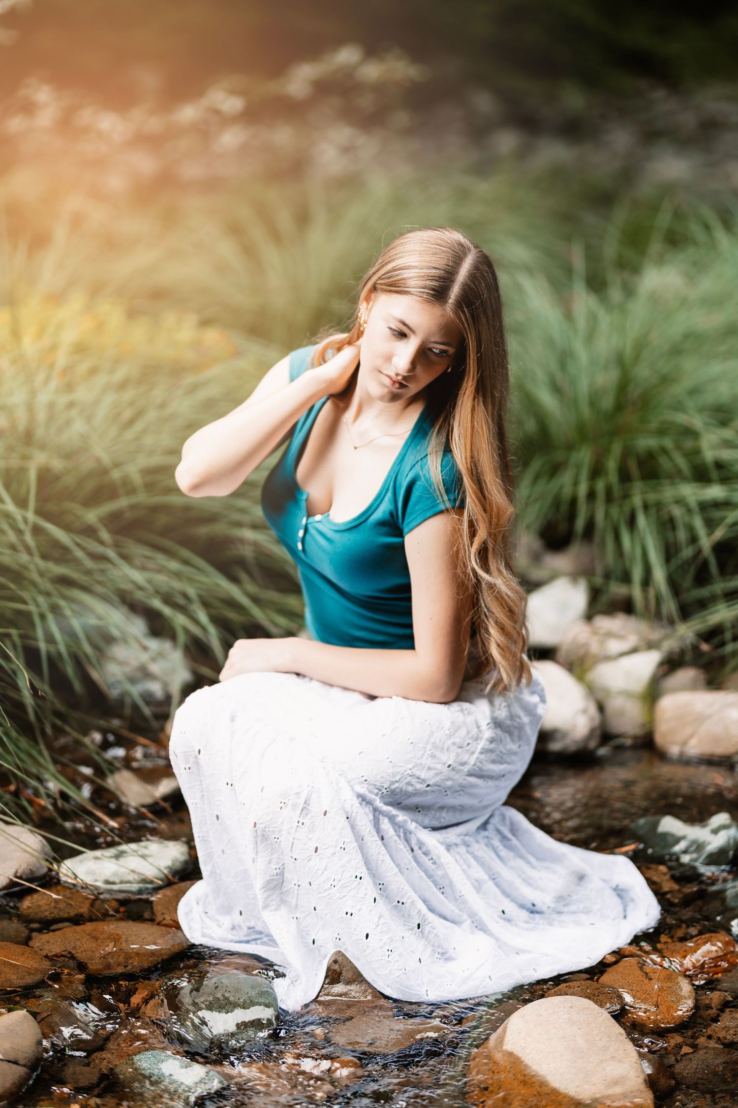 Young woman with long wavy hair in a teal top and white skirt sitting on rocks by a stream.