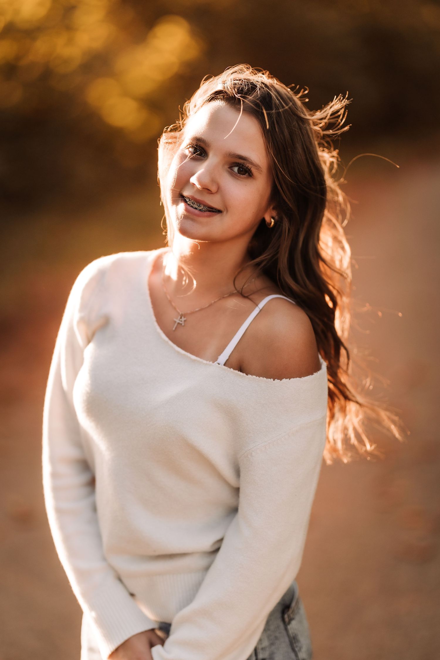Smiling brunette teen girl with braces in white off-shoulder sweater posing in warm autumn sunlight.