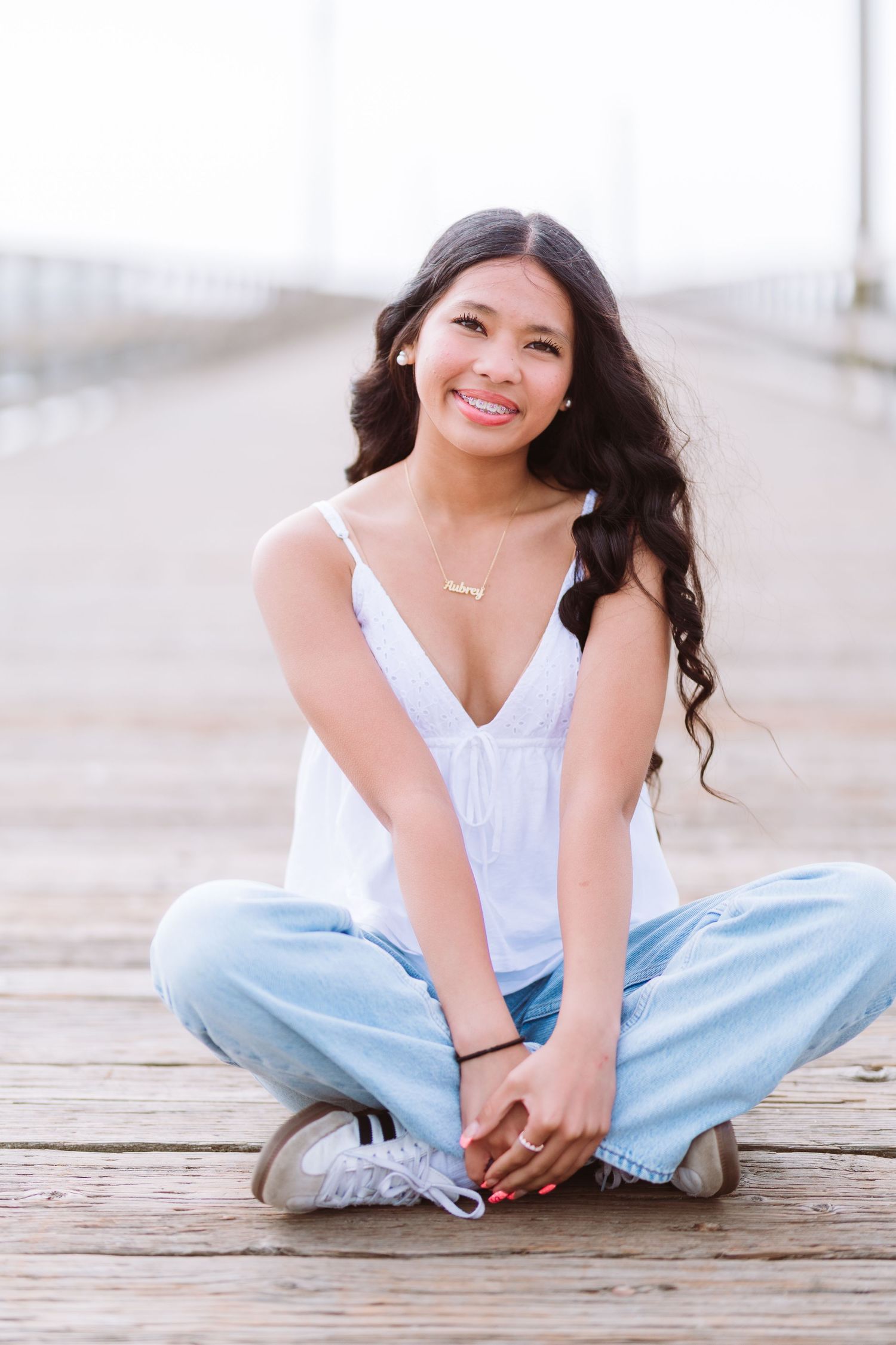 Young woman with long dark hair sits cross-legged on wooden pier, wearing white top and light jeans, smiling warmly.