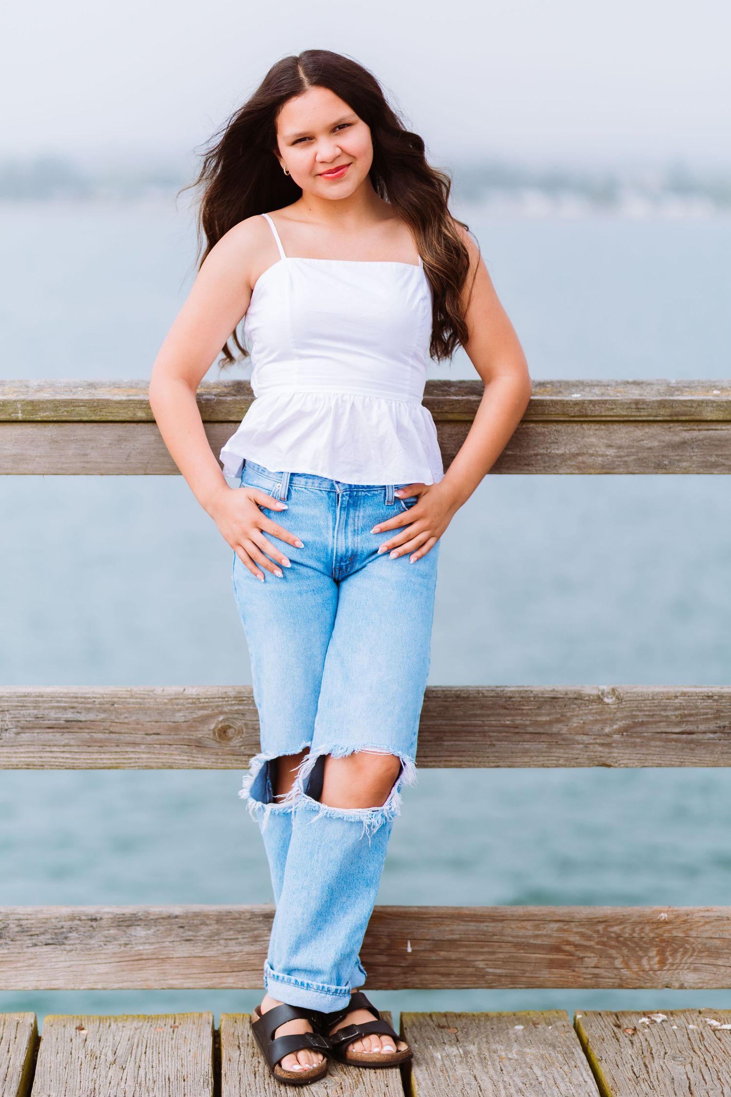 Young woman in white top and ripped jeans posing on wooden pier with water in background.