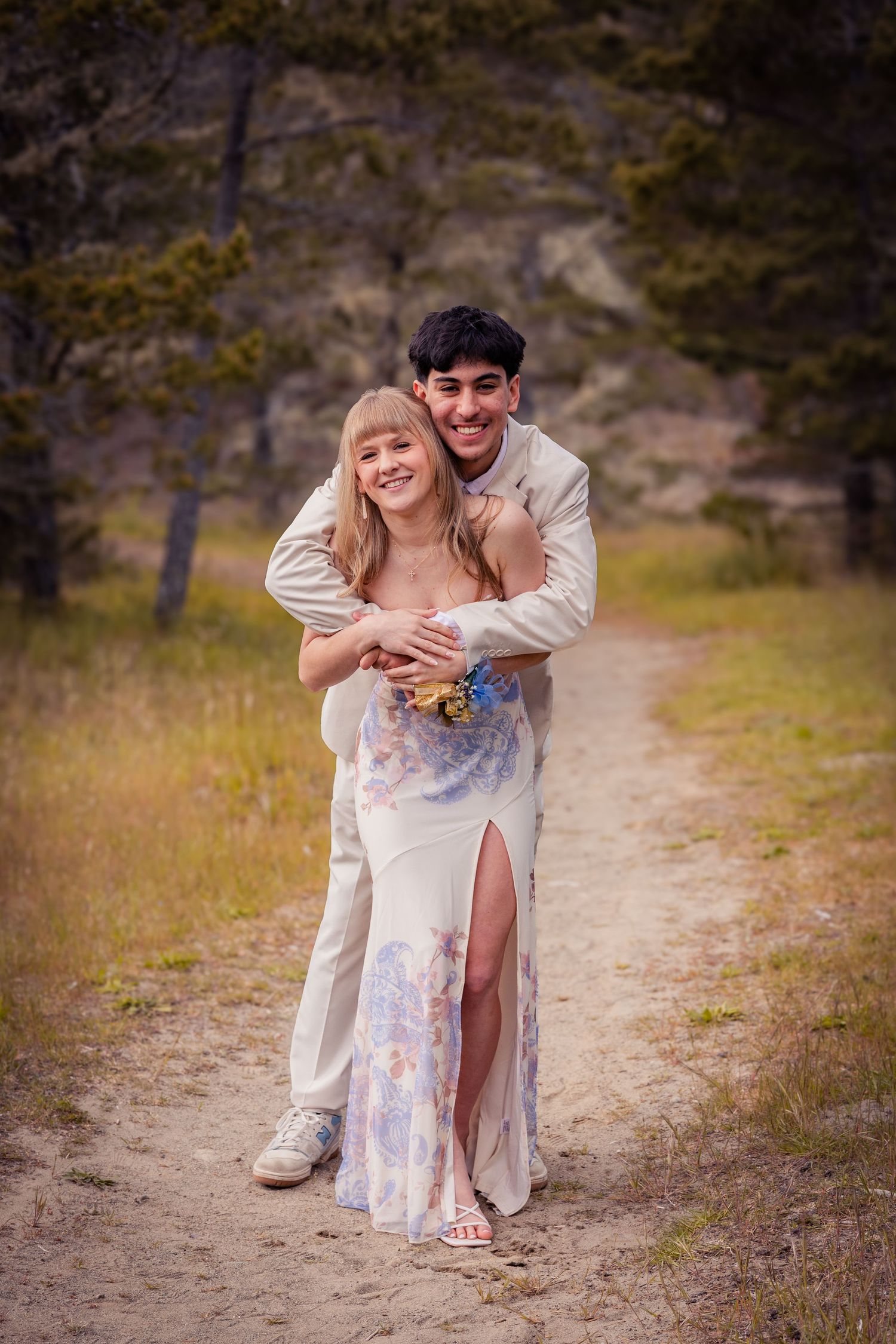 A smiling couple in prom attire hugs playfully on a dirt path with a scenic natural backdrop.