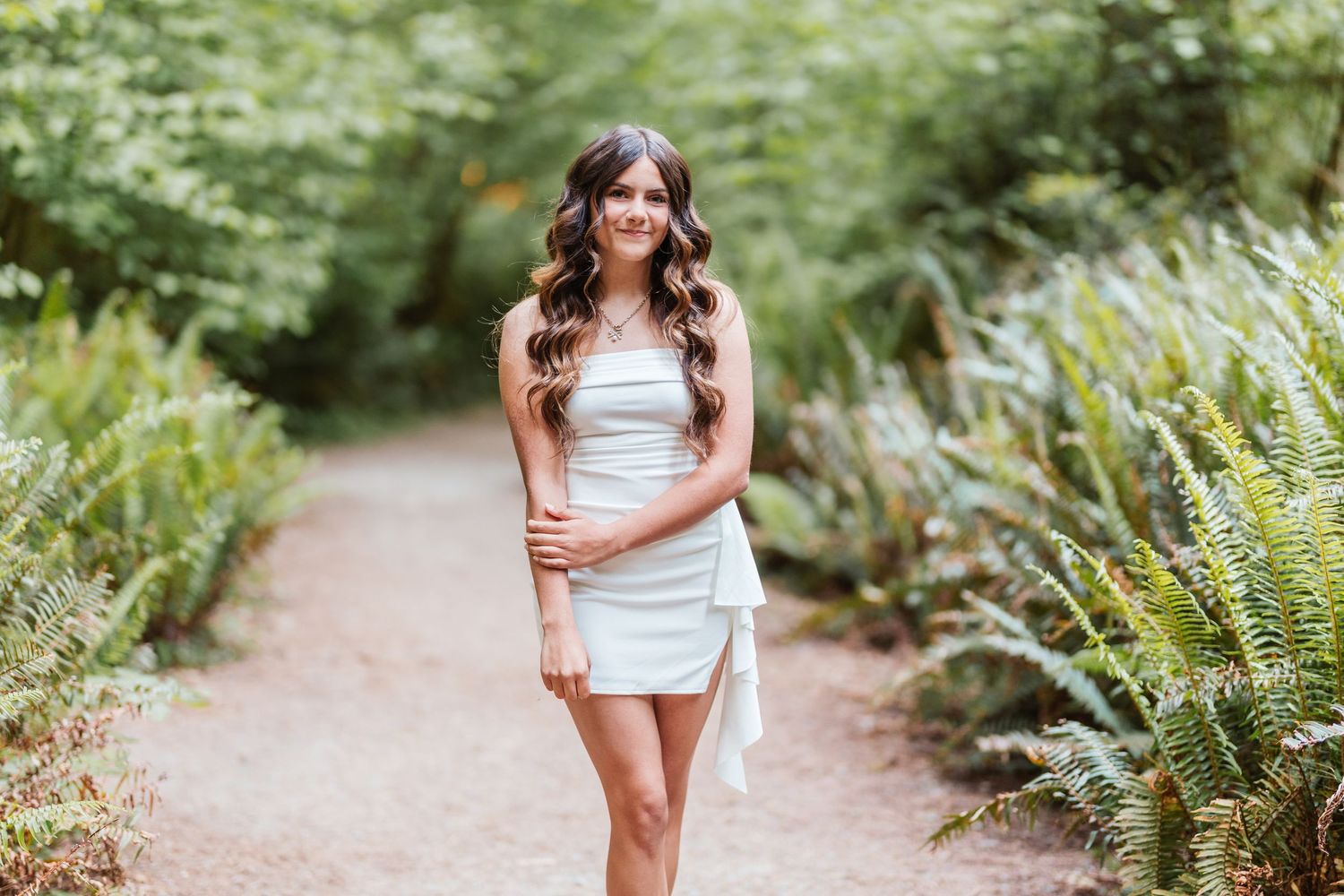 Young woman in white mini dress walking on a garden path surrounded by lush green ferns and trees.