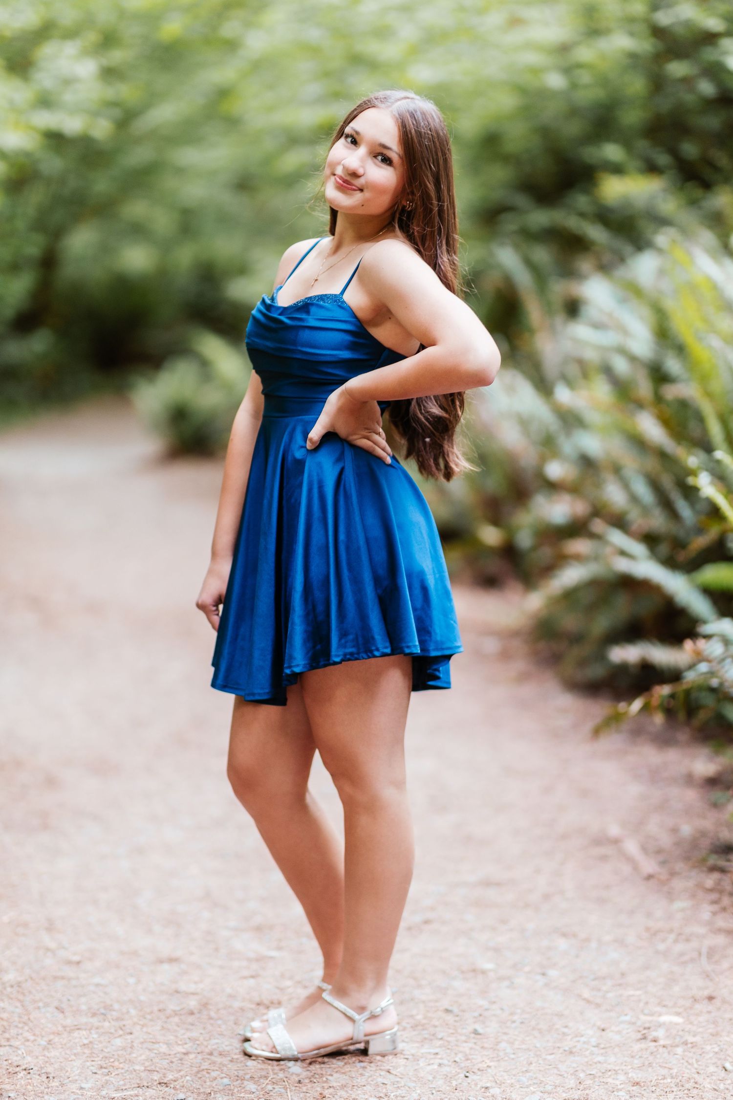 Young woman in blue satin mini dress standing on garden path with hand on hip, wearing silver flats.