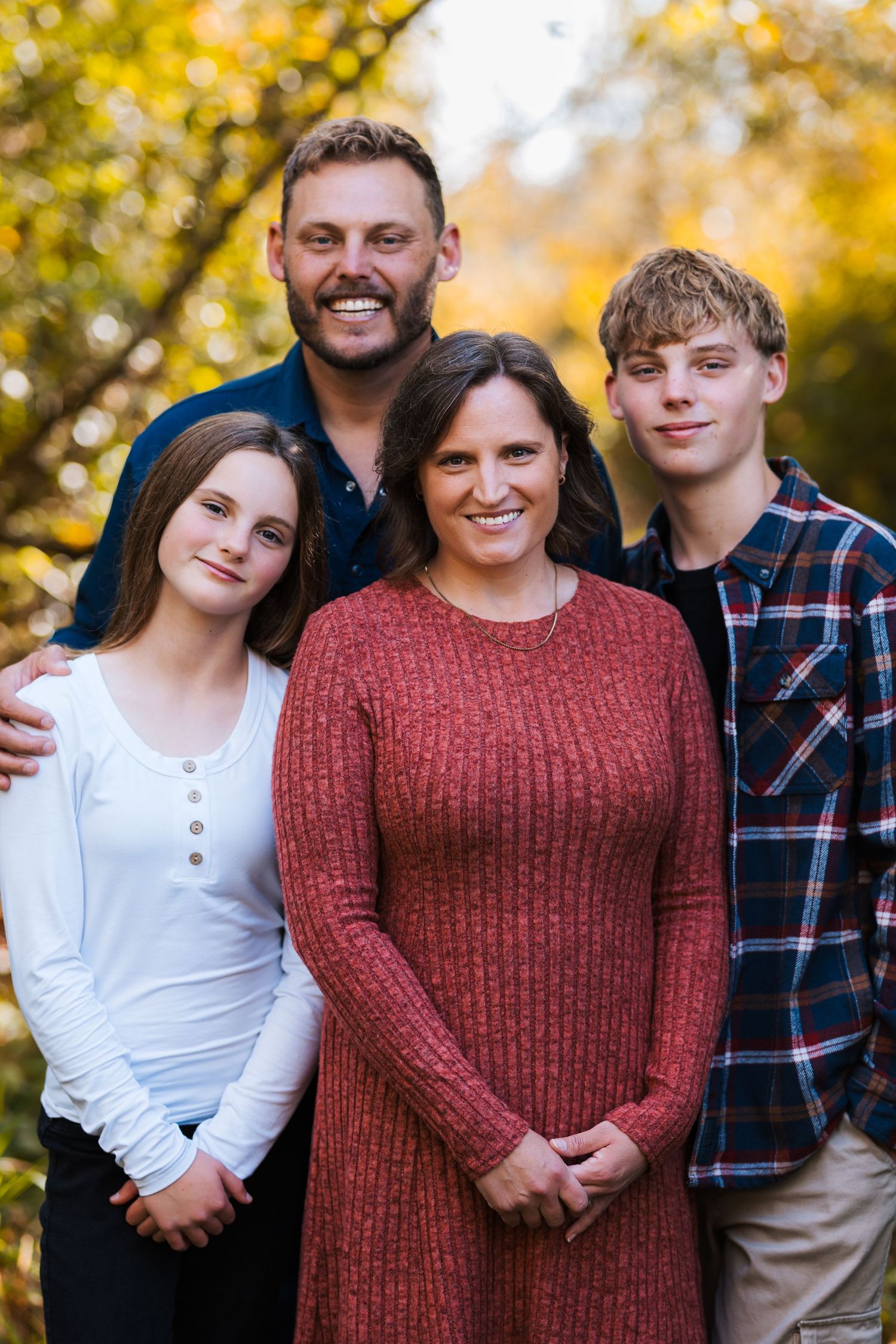 Family of four smiling outdoors in autumn, surrounded by golden foliage during a fall portrait session.