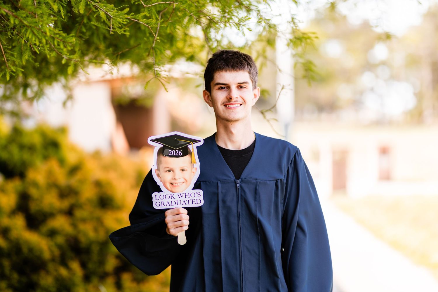 Young man in blue graduation gown holding a grad cap photo prop outdoors, smiling proudly on graduation day.