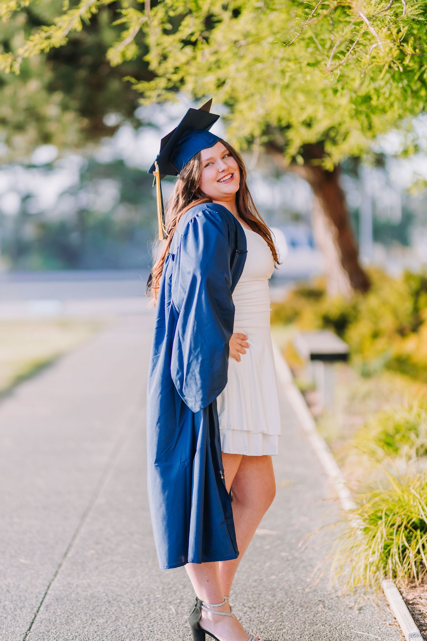 Smiling graduate in blue cap and gown poses outdoors on a sunny day wearing a white dress underneath.