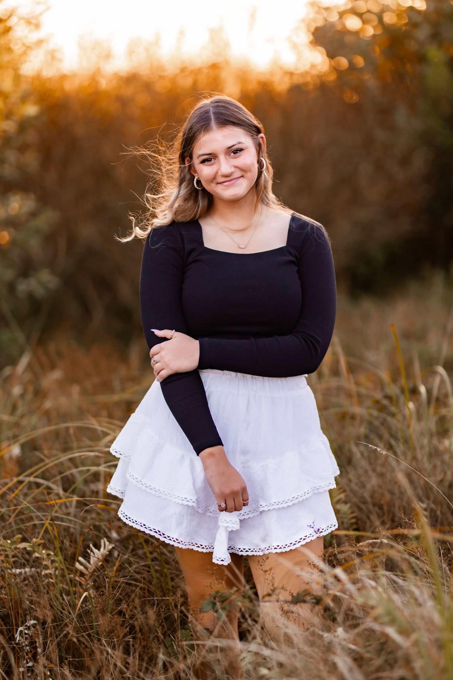 Smiling blonde teen girl in black long-sleeve top and white tiered skirt stands in autumn field.