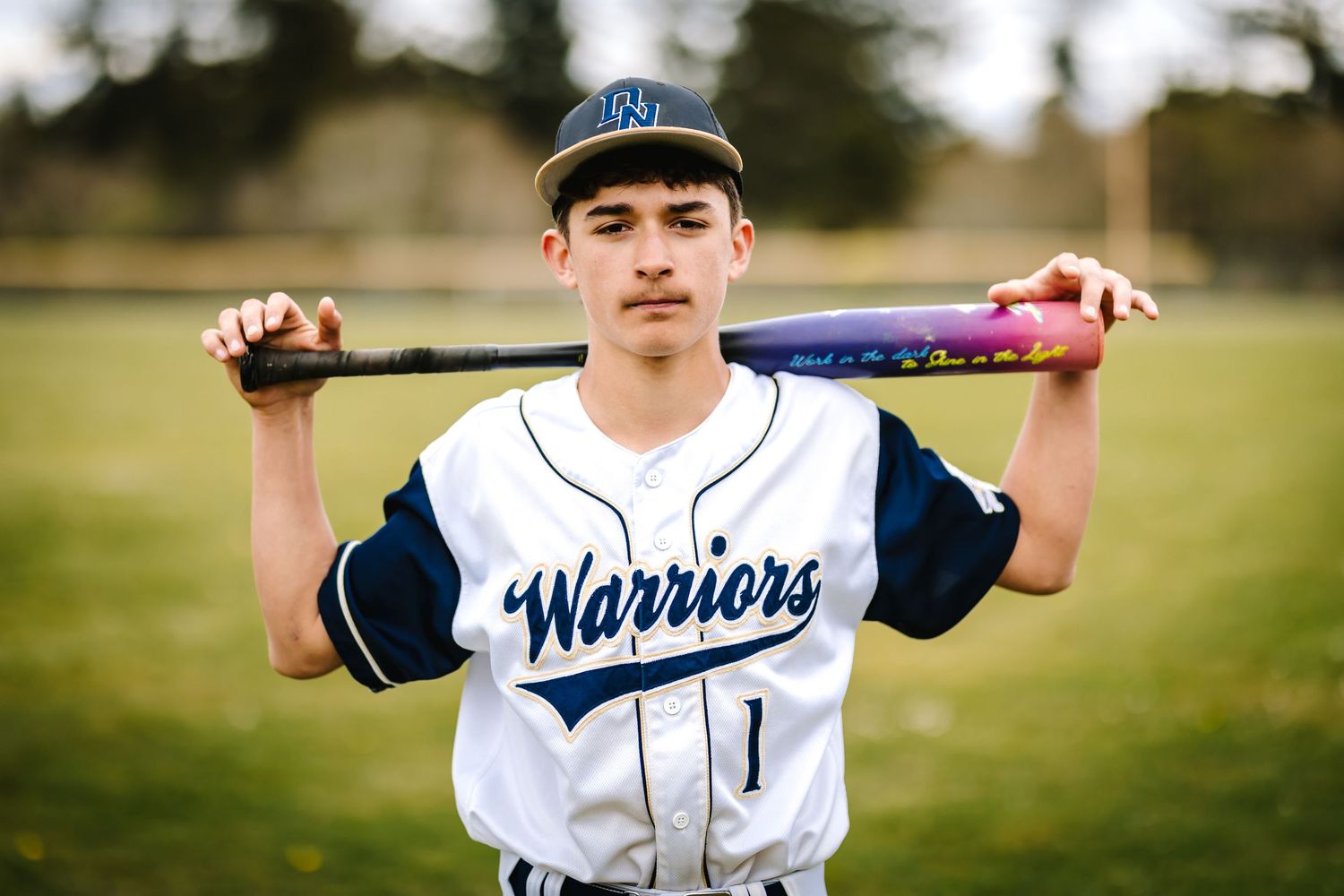 Young male baseball player in Warriors uniform number 1 poses with bat across shoulders on a field.