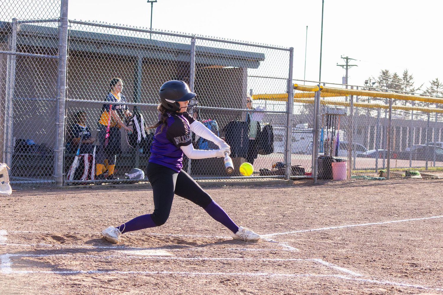 Female softball player in purple uniform makes contact with ball during a game on a dirt infield.