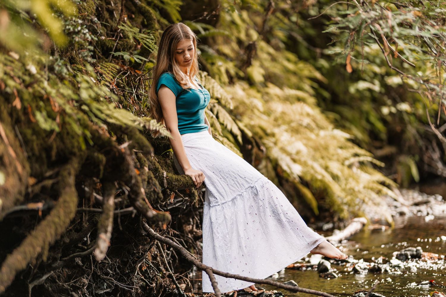 Young woman in teal top and white maxi skirt exploring a mossy forest creek.