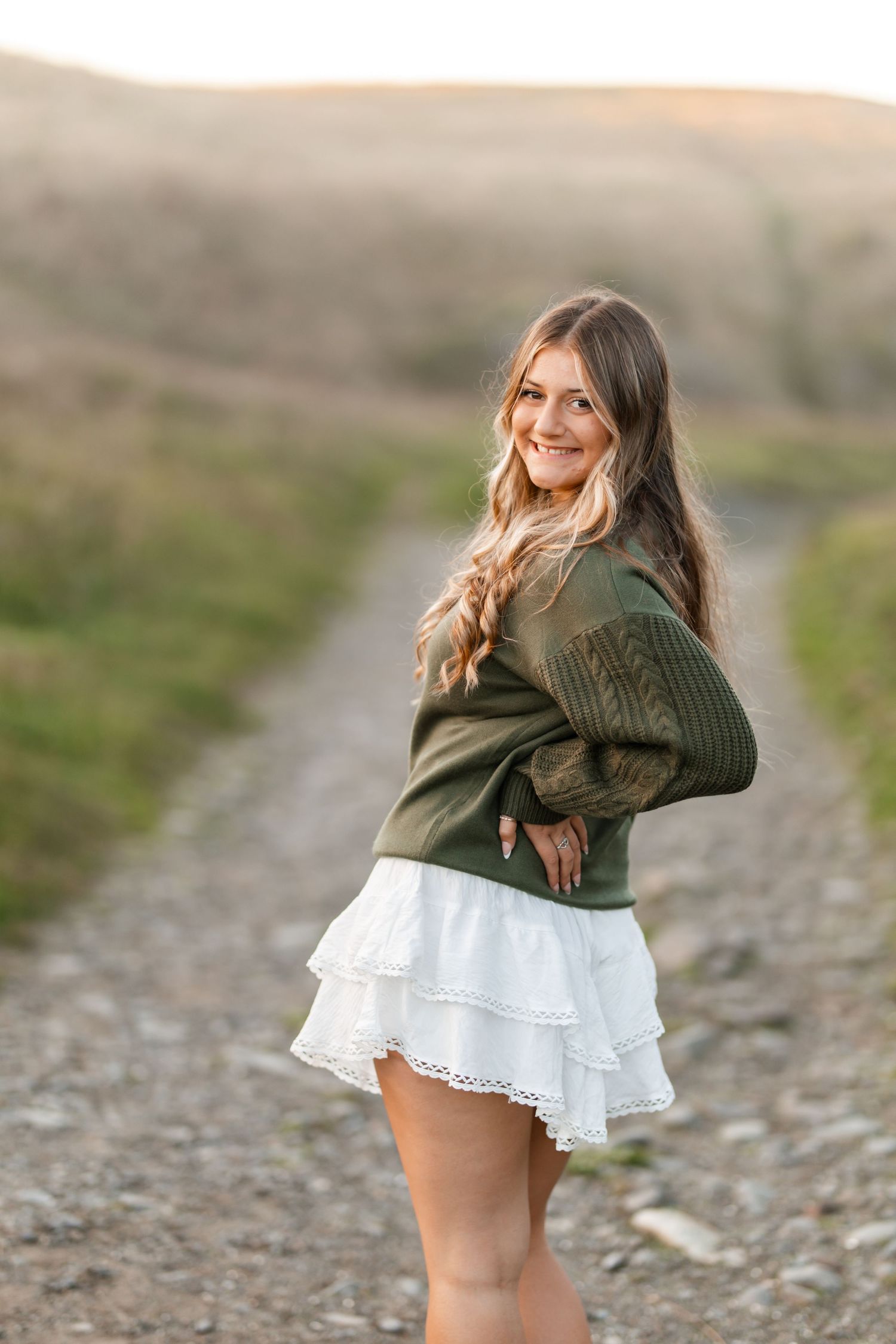 Young woman in green knit sweater and white ruffle skirt posing on a sunlit gravel path outdoors.