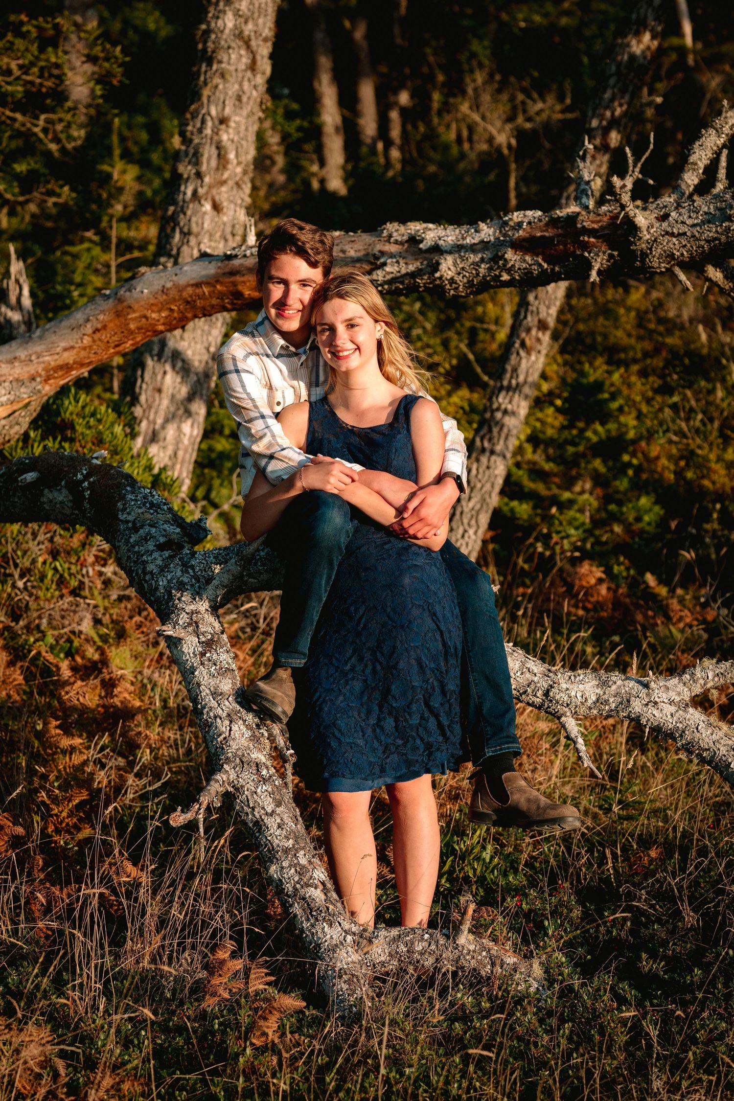 A smiling couple posing on a fallen tree in a forest, woman in blue dress with man behind her.