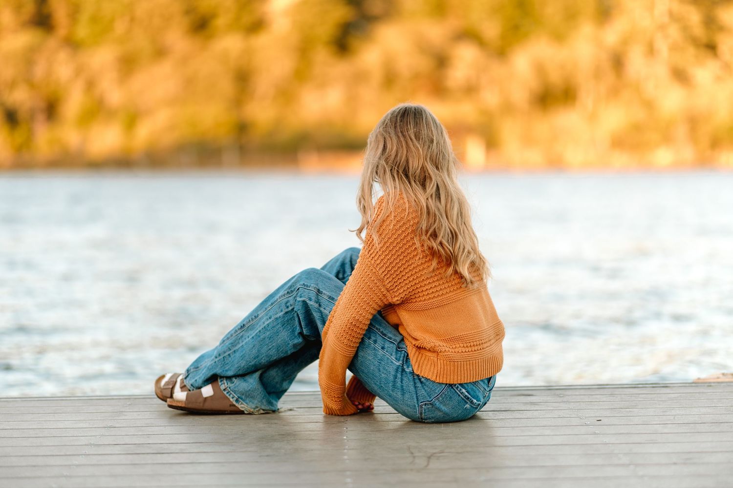 Blonde girl in orange sweater and jeans sitting on a dock gazing at a calm lake in autumn.