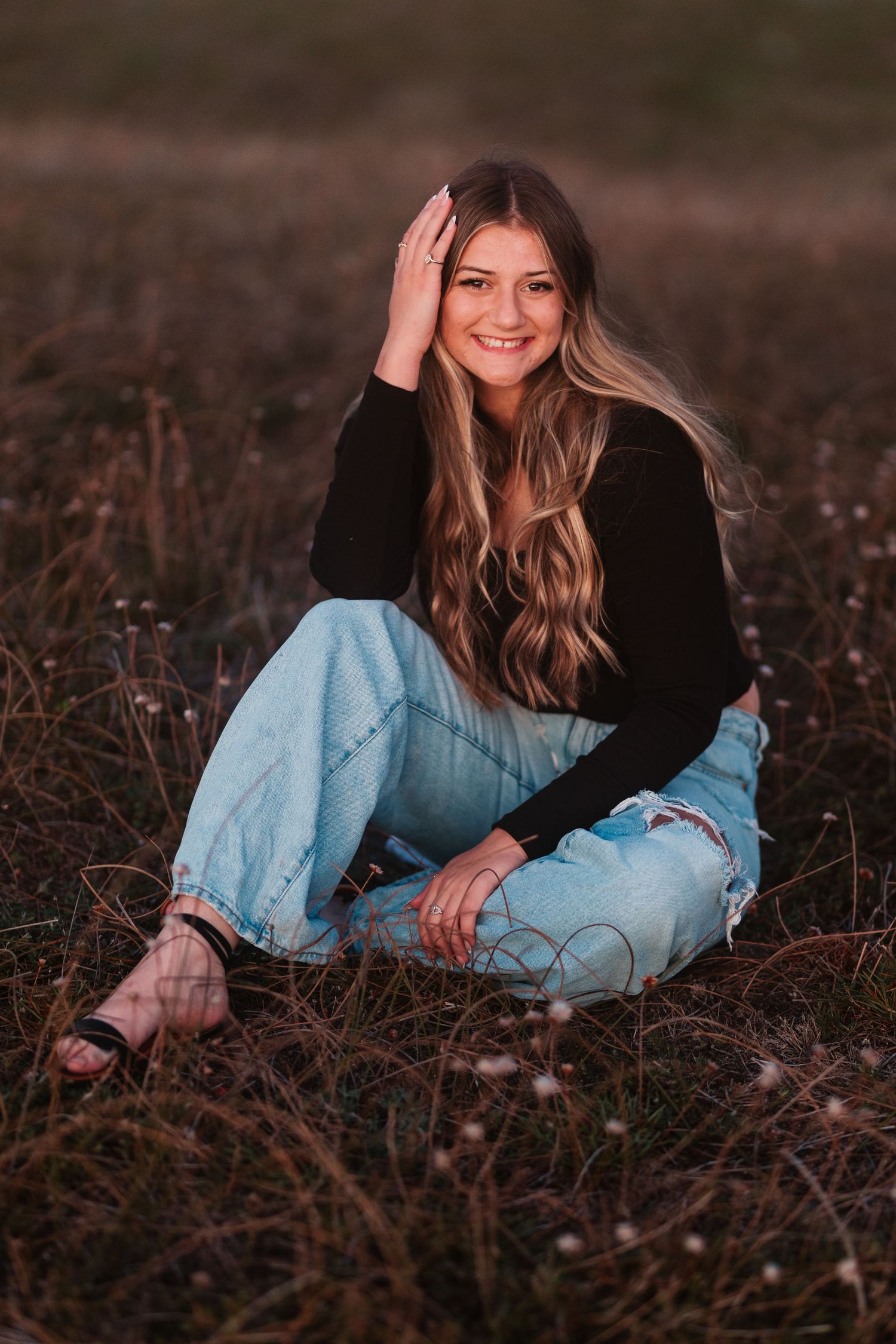 Smiling teenage girl in black top and ripped jeans sitting in golden field at sunset.