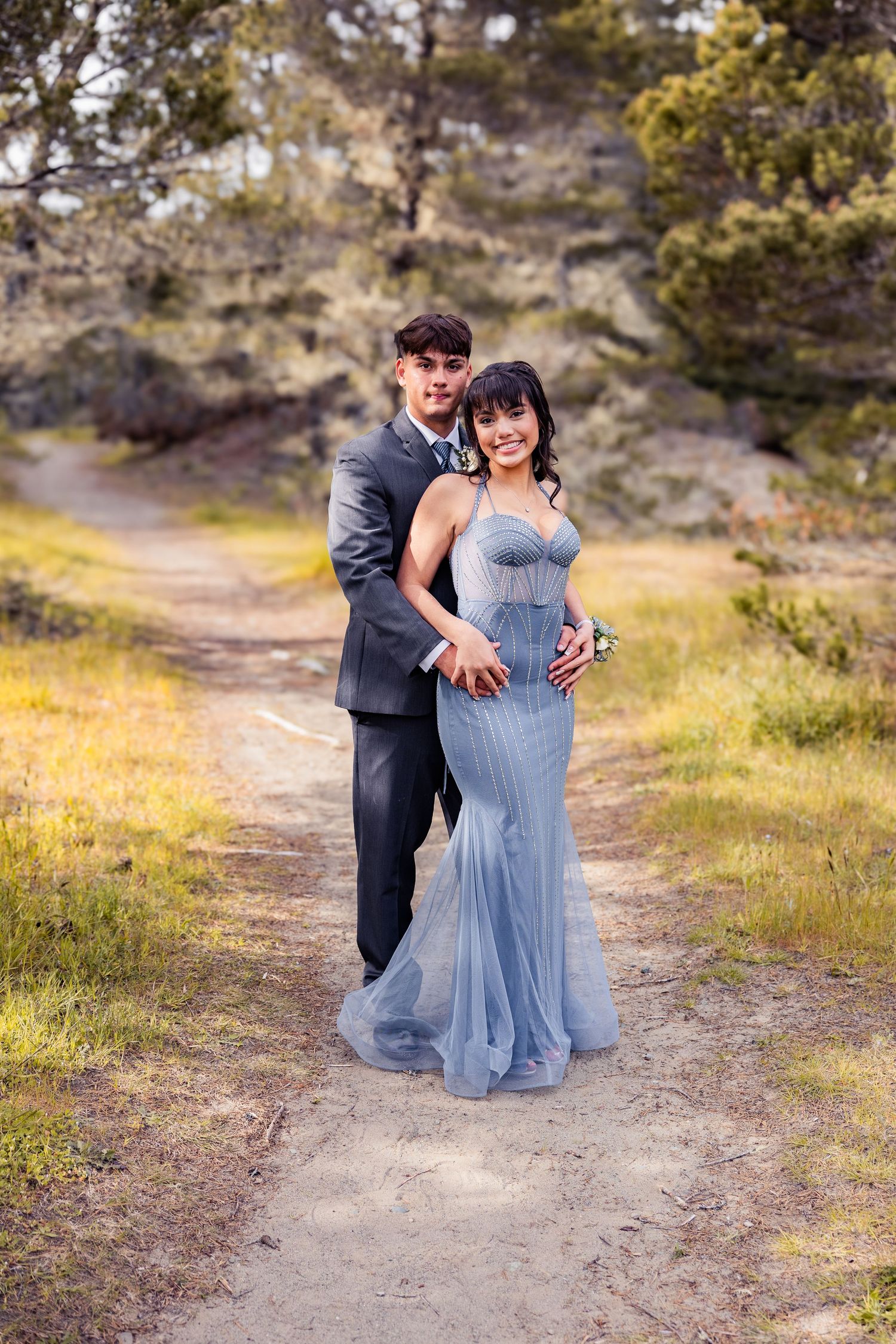 A couple in formal prom attire poses on a nature trail, the woman in a blue gown and man in a gray suit.